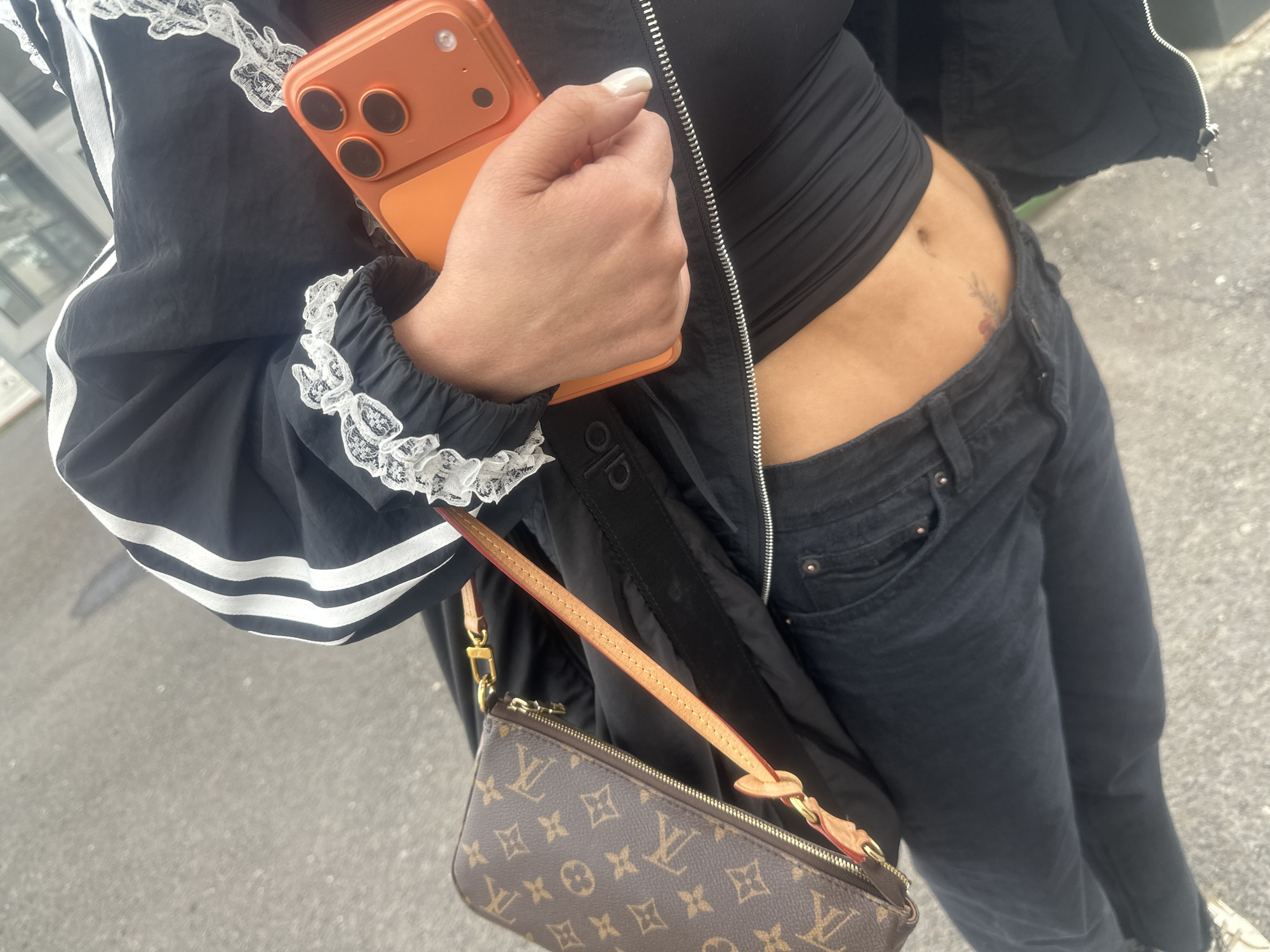 A person stands in an outdoor urban environment, likely in the city of Yerevan, Armenia. The person wears black jeans, a black top, and a black jacket with white stripes and lace cuffs. They are holding an orange smartphone in their left hand. A brown Louis Vuitton Pochette Accessoires handbag hangs from their shoulder. A small floral tattoo is visible on the person's stomach. The background appears to be a city street with a sidewalk.