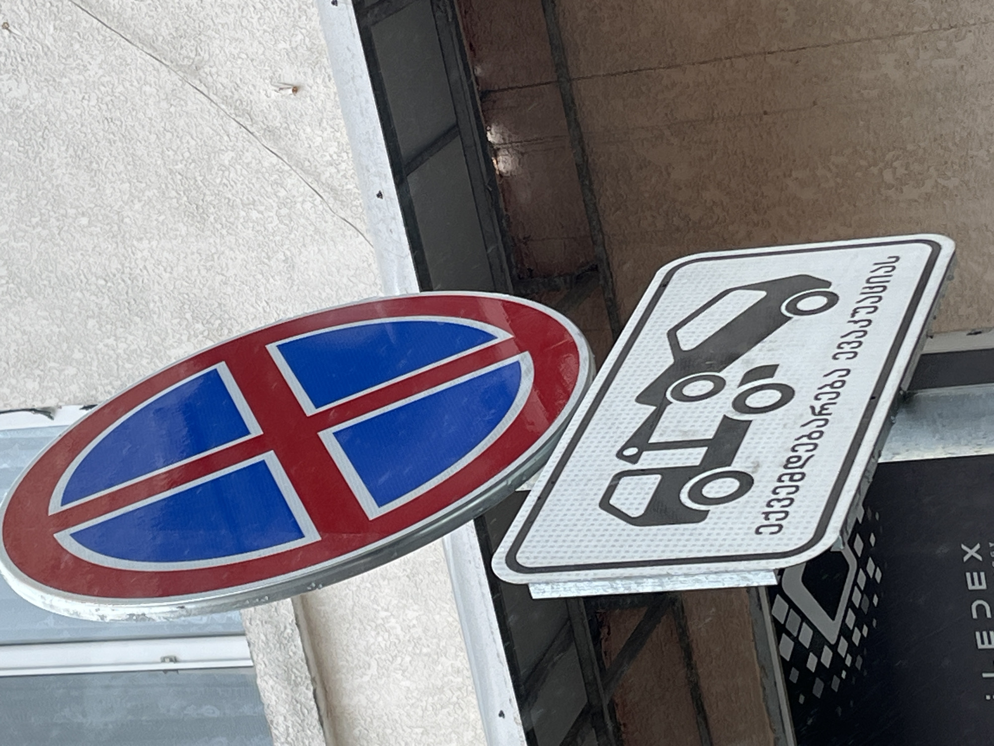 The image shows two road signs mounted on a pole. The upper sign is a red and blue oval sign with a red cross, indicating no stopping. Below it, a white rectangular sign displays a black icon of a tow truck lifting a car. This sign also has text in Georgian script, which translates to "Towing service". The signs are positioned in front of a building with a textured light-colored facade and a metal overhang. The overall scene suggests a street in Batumi, Georgia, where parking restrictions are enforced by towing.