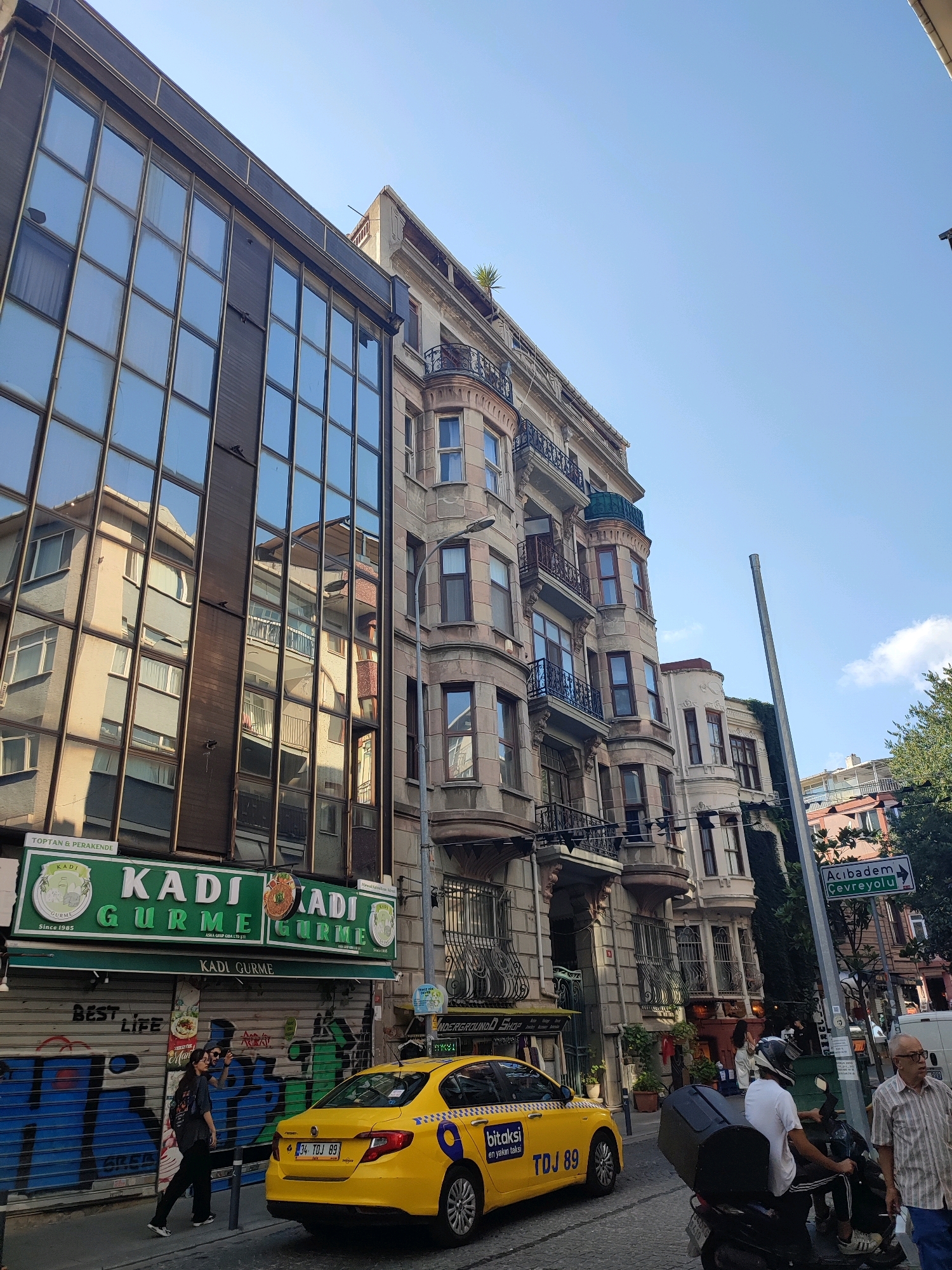 This is a street scene in Kadıköy, Turkey, on a clear day. A modern building with large, reflective windows stands to the left, mirroring the older, ornate architecture of the building next to it. The older building features multiple balconies with intricate iron railings, bay windows, and decorative architectural details.
In the foreground, a yellow taxi with the license plate "34 TDJ 89" is parked on the street. A motorcycle with a rider wearing a white shirt and helmet is next to the taxi. Two pedestrians are walking on the sidewalk in front of a shop with a green awning that reads "KADI GURME". The shop's roll-down security gate is partially covered in graffiti.
Further down the street, a green directional sign points towards "Acıbadem Çevre Yolu". Trees are visible on the right side of the image, and the sky is a bright, clear blue. The overall impression is a bustling urban environment with a mix of old and new architecture.