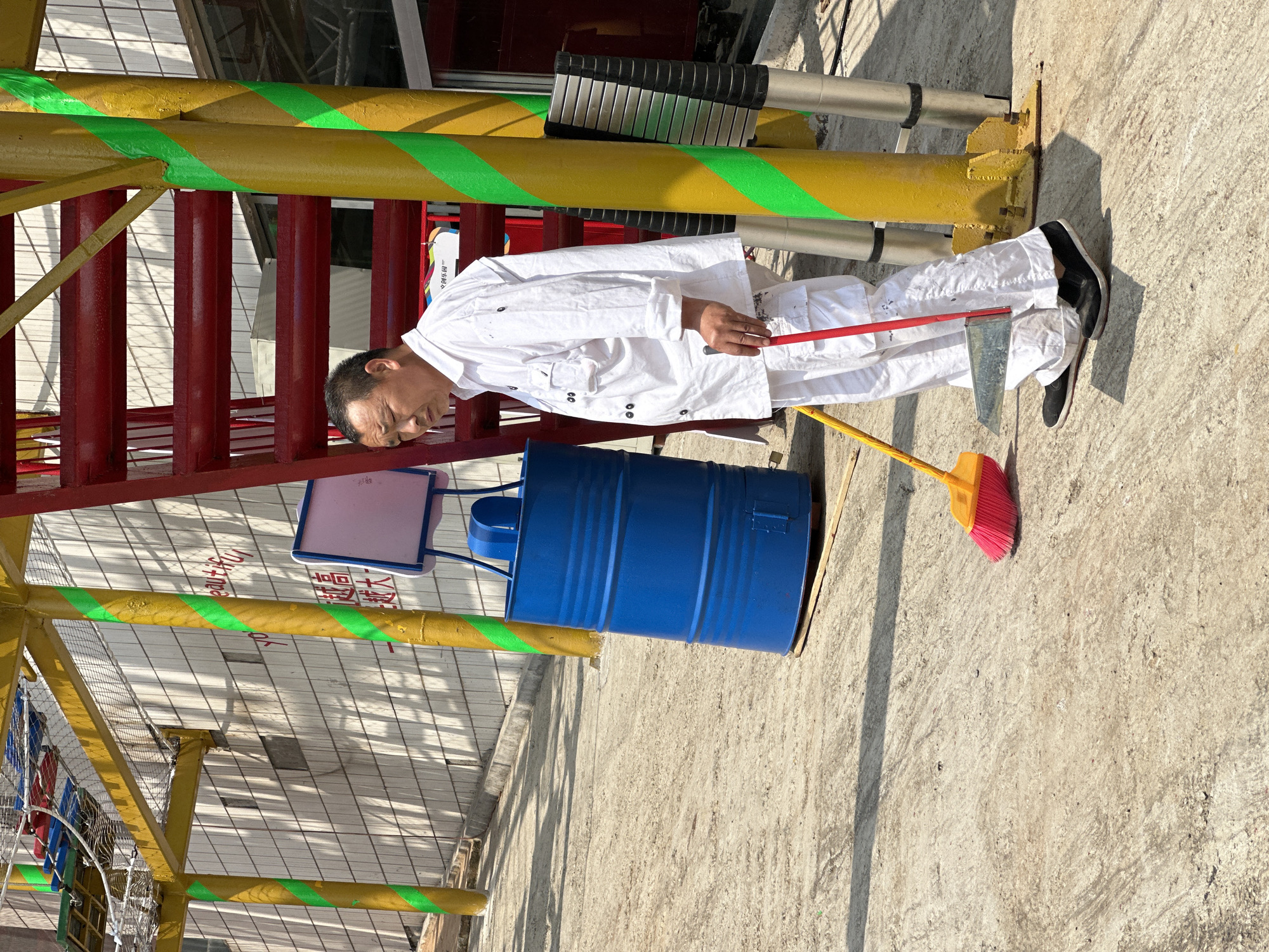 The scene depicts a man standing on a concrete surface near an outdoor playground structure. The man is wearing a white jacket and pants with black shoes. He has a red-handled squeegee in his right hand and is standing near a blue metal barrel. A yellow broom with a red head is next to the barrel. The playground structure includes red, yellow, and green painted elements. The location is in Nan'an District, China.