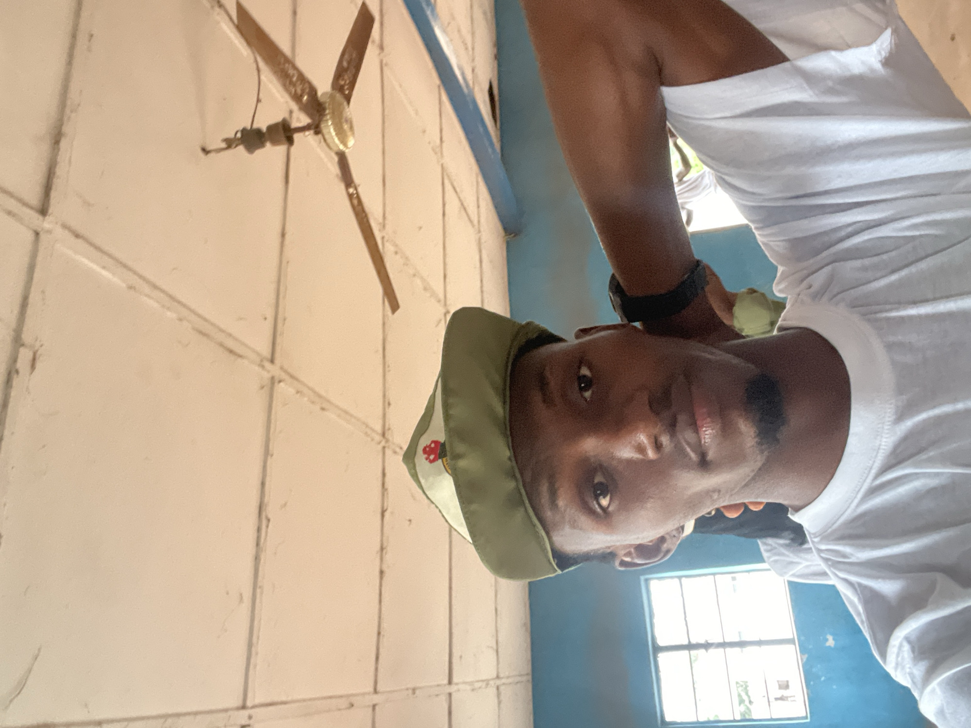 A young Black man wearing a green cap and a white t-shirt takes a selfie indoors. The man has short black hair, brown eyes, and a goatee. He is looking directly at the camera with a neutral expression. His left arm is bent behind his head, and he is wearing a black watch on his left wrist. In the background, there is a white ceiling with a fan, a blue wall, and a window with bars. The scene appears to be a classroom or a simple room.