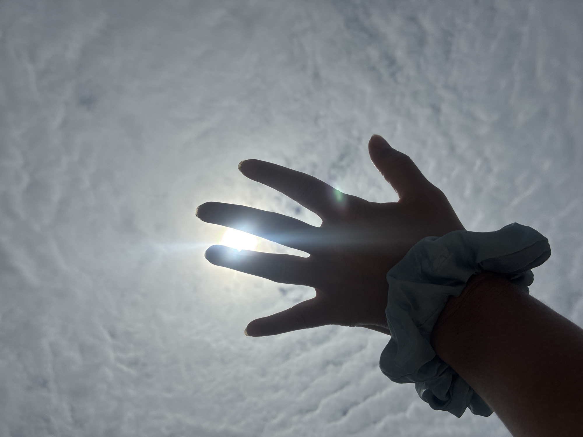 A hand with a blue scrunchie on the wrist is silhouetted against a bright sky. The sun is visible through the fingers, creating a lens flare. The sky is filled with wispy clouds. The scene appears to be outdoors, likely in a city or urban setting, but the specific location of Sochi, Russia cannot be determined from the image alone.