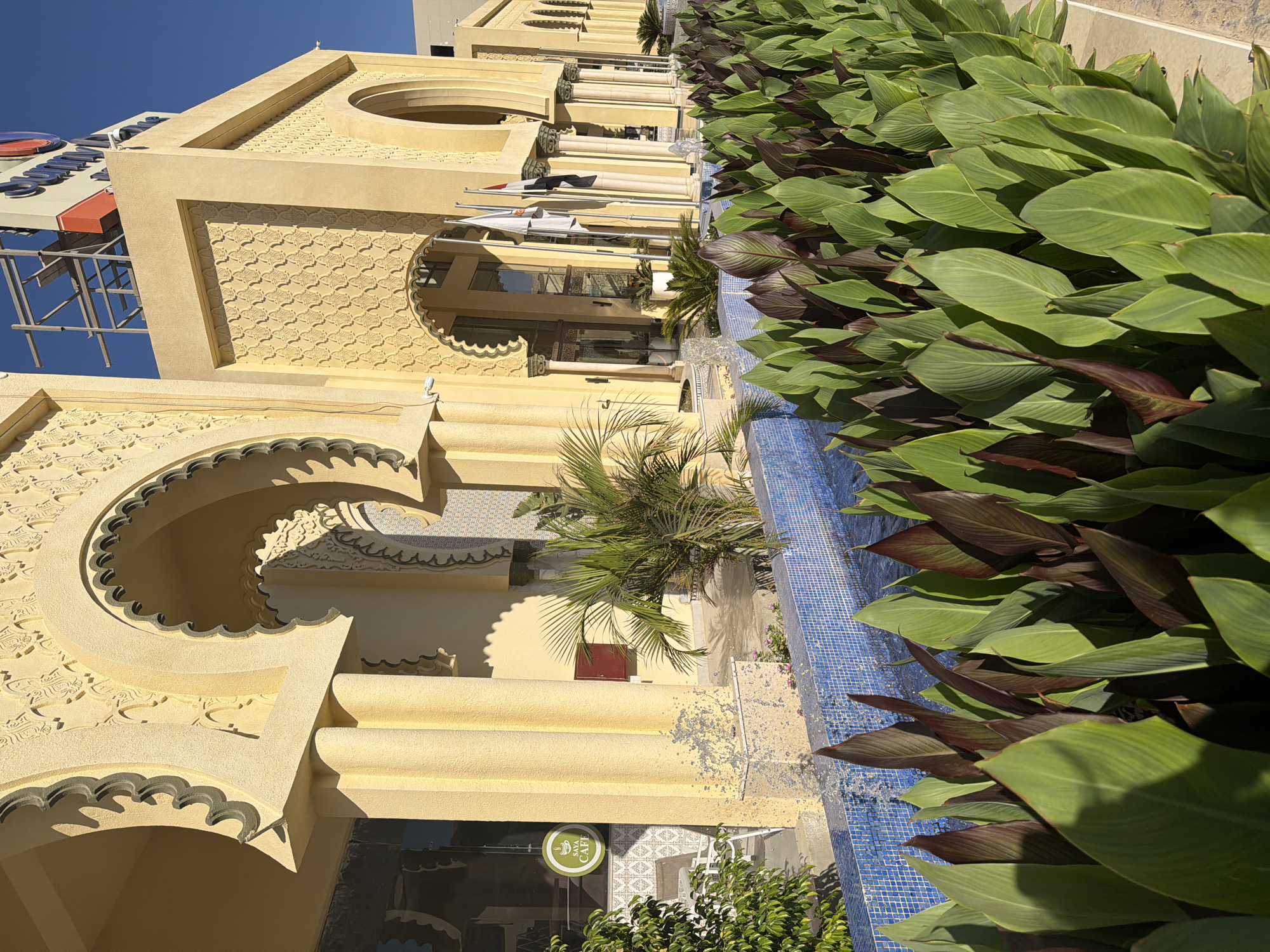 The image shows a sunny day in Hurghada, Egypt, with a focus on the exterior of a building featuring traditional Islamic architecture. The building is painted in a warm, sandy yellow and is adorned with intricate decorative carvings and arches. A row of lush green plants with deep red undersides lines a bright blue tiled water feature in the foreground.
In the background, a tall building and a large sign with red and blue lettering are visible against a clear blue sky. The architectural style of the main building, with its decorative patterns and arched entrances, suggests a commercial or hospitality establishment, possibly a hotel or a shopping complex. The presence of palm trees adds to the tropical ambiance.
The lighting indicates it is daytime, with strong sunlight casting distinct shadows. The scene appears to be peaceful and well-maintained, suggesting a resort or leisure location. No people are visible in the image, but the presence of outdoor seating and the "Sava Cafe" sign indicates a place where people would gather. The overall impression is one of a pleasant, warm, and visually appealing environment typical of a resort town in Egypt.