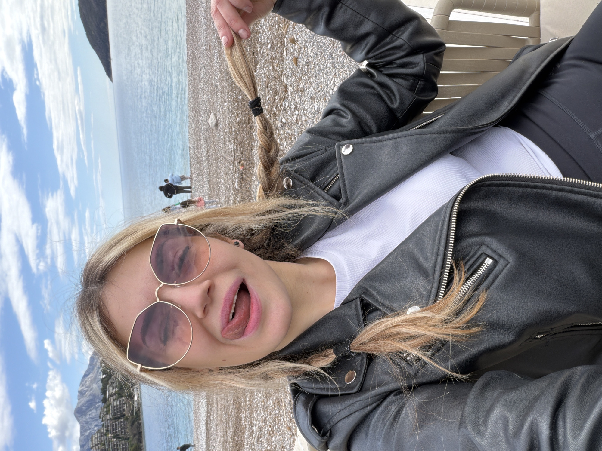 The image depicts a woman with blonde hair in pigtails, wearing rose-tinted sunglasses, a white shirt, and a black leather jacket. She is sticking out her tongue, and holding one of her pigtails. She is sitting on a white plastic chair on a beach, likely in Budva, Montenegro, based on the mountain and coastline in the background. A few people are visible in the distance along the shoreline. The weather appears sunny with a partly cloudy sky.
