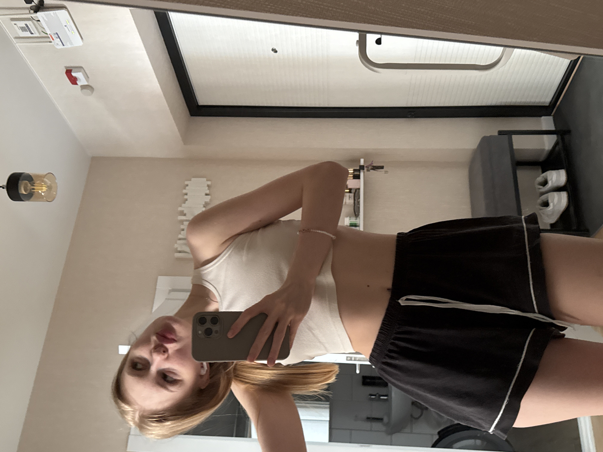 A young woman is taking a selfie in a bathroom. The woman is wearing a white tank top and black shorts with a white trim. She is holding a phone in her right hand. The bathroom has a white sink and a black shower door frame. There are tiles on the wall behind her. A light fixture with an exposed bulb is visible. A shelf with shoes can be seen in the reflection. The location is in a city, likely in Perm, Russia based on the fixtures and style of the room.