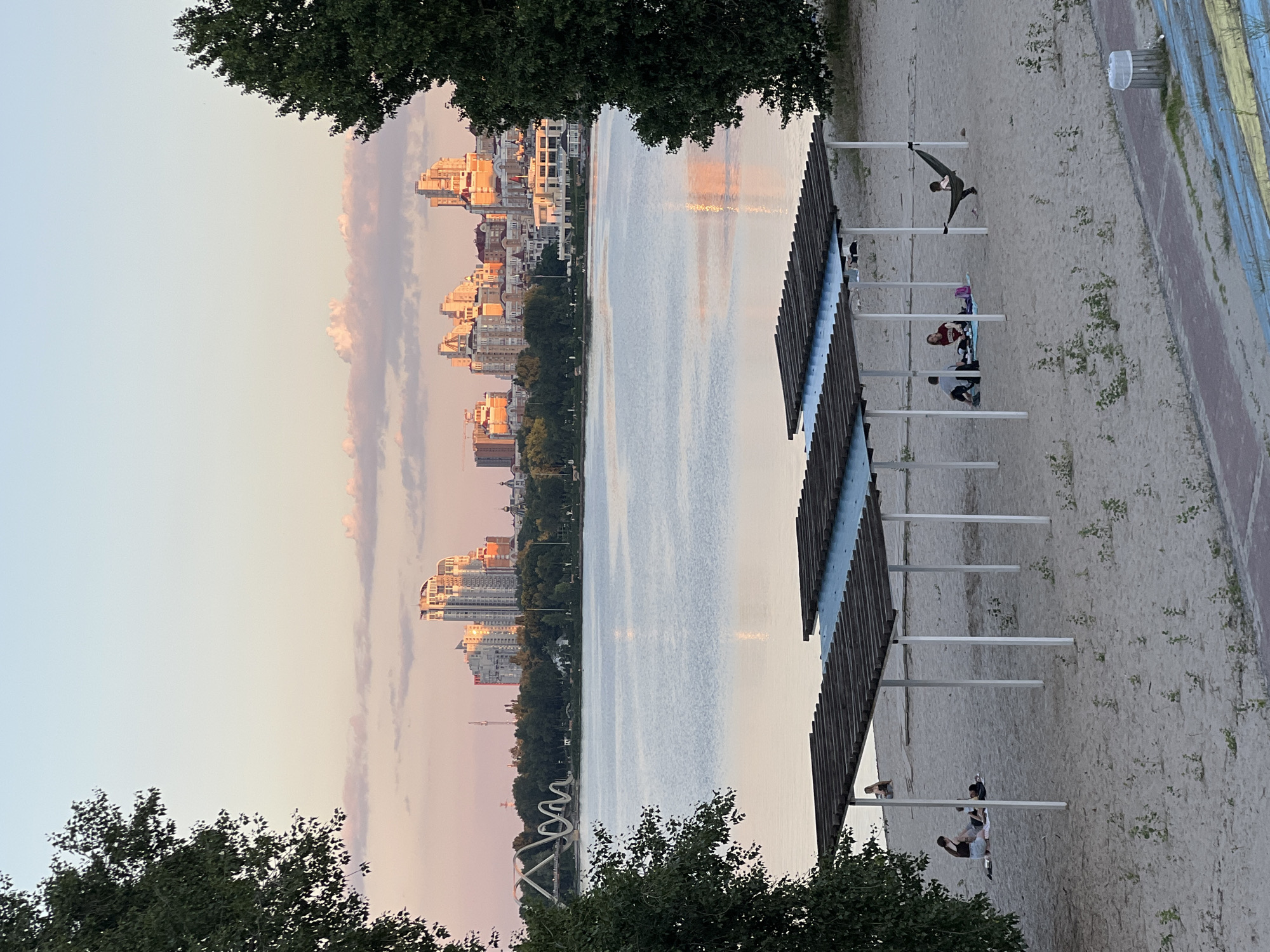 This is a daytime, wide-angle, eye-level shot of a beach scene in Kyiv, Ukraine, with a city skyline and river in the background. The foreground features a sandy beach with several wooden canopies supported by white poles, providing shade. Underneath these canopies, various individuals are engaged in leisure activities.
On the left, under the first canopy, two people are seated on a blanket, seemingly having a conversation. To their right, a couple is lying down on a blanket, with one person appearing to read. Further to the right, under the second canopy, a group of three individuals are gathered, with one person lying in a hammock attached to one of the canopy poles. Another person is visible sitting in the distance under the furthest canopy on the left.
The beach itself is a light tan sand with scattered small green plants. A paved pathway runs along the bottom right of the image, marked with colored stripes. Beyond the beach is a wide, calm river, reflecting the soft hues of the sky. In the distance, across the river, lies the Kyiv cityscape. Tall, modern buildings with light-colored facades dominate the skyline, bathed in the warm glow of sunset. A distinctive white, arching bridge is visible to the left of the buildings. Lush green trees line the riverbank in front of the city. The sky is a clear pale blue, with scattered, soft pink and white clouds. The overall atmosphere is one of peaceful relaxation in a scenic urban setting.