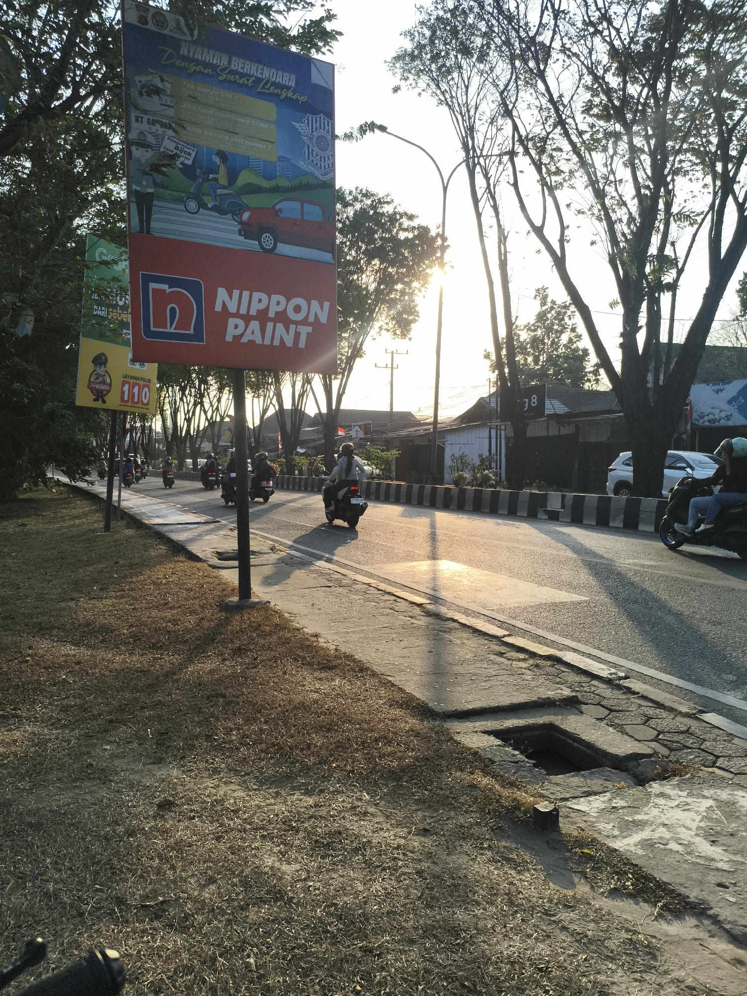 The image shows a street scene in Samarinda, Indonesia, during the golden hour. The sun is low in the sky, casting long shadows and illuminating the road and surrounding trees. A large billboard for Nippon Paint is prominent on the left side of the image. The billboard features an advertisement with cartoonish illustrations and text in Indonesian, promoting safe driving with complete documents.
The street is paved with asphalt and bordered by a concrete sidewalk. A concrete drainage grate is visible on the sidewalk, slightly damaged. The grass on the side of the road is dry and appears to be unkempt. Several motorcycles are riding on the road, with riders wearing helmets. One motorcycle in the foreground has two passengers. In the background, there are buildings and more trees. The overall impression is a typical urban street scene in Indonesia during sunset.