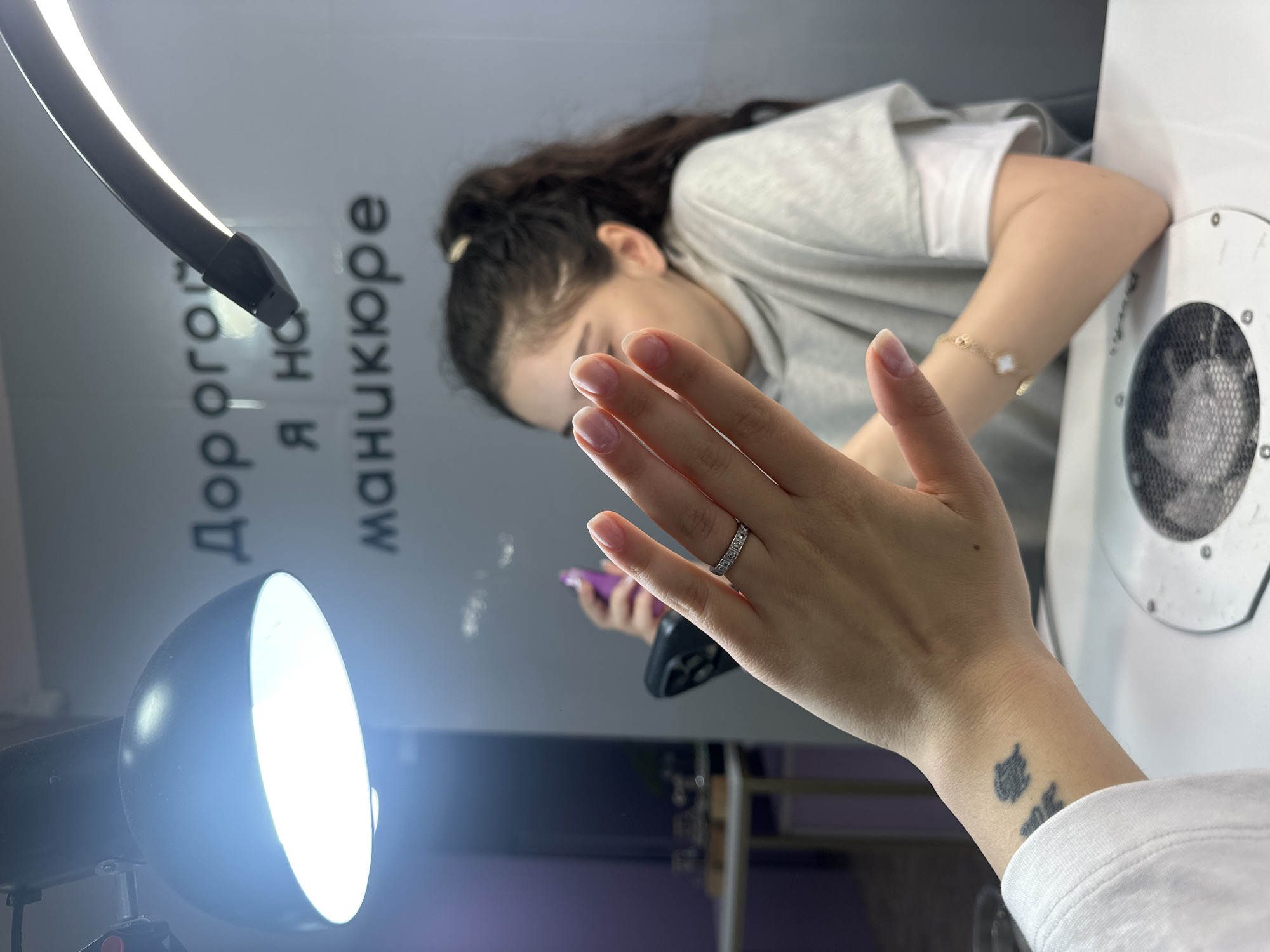A young woman with long, dark hair pulled back in a ponytail is receiving a manicure. She is wearing a gray t-shirt and has a gold bracelet on her right wrist. On her left hand, she wears a ring on her finger, and there is a small tattoo on her left wrist. She holds a purple phone in her left hand. Her right hand is outstretched, showing her fingernails. A ring light and a round lamp on a tripod provide illumination. Text is visible on the wall in Cyrillic script: "Дорого я на маникюре". The scene is likely indoors in a salon, potentially in Yerevan, Armenia, based on the Cyrillic text and image characteristics. There is a manicure machine with a fan on a white table on the right.