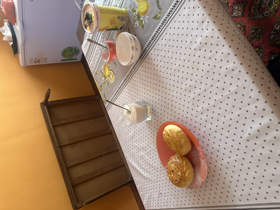 A table is set with a white and gray polka-dot tablecloth. A container of food, a glass of pink liquid with a spoon, and a white bowl are in the top left corner. Further down is a glass filled with sugar and a spoon, next to a plate with two buns, one with sesame seeds. A wooden frame is attached to the wall on the left side of the image.