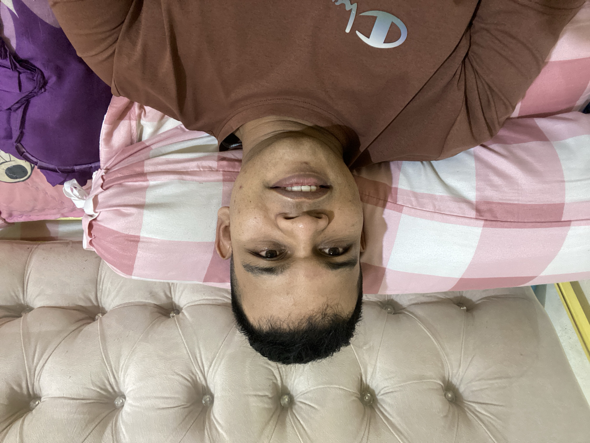 A medium shot shows a man lying on a bed, looking directly at the camera. He has short, dark hair and brown eyes. He is wearing a brown t-shirt with a white logo on the left chest. He is propped up against a light-colored, tufted headboard. In front of him is a pink and white checkered pillow. To his right, partially visible, is a purple pillow with a cartoon character on it. The scene appears to be indoors, likely a bedroom in Lambaro Kueh, Indonesia.