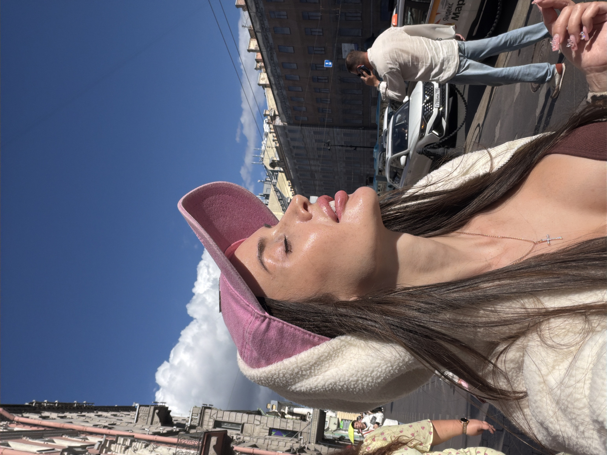 The media file shows a young woman with long, dark hair, wearing a pink cap and a white, fuzzy jacket, against a backdrop of buildings and a bright blue sky with fluffy white clouds. She is looking upwards, with her eyes closed and her mouth slightly open, appearing to be basking in the sun. A delicate silver necklace with a cross pendant is visible around her neck. The scene is an urban environment, with a street, cars, and buildings that suggest a city setting. In the background, a man is walking and talking on a cell phone. One of the cars has a license plate that reads "786 AA 196". A white van in the background has text on its side, including "Яндекс Маркет". The time of day appears to be daytime, likely midday, given the bright sunlight casting shadows and highlighting the woman's face. The weather seems to be clear and sunny. The overall mood is relaxed and perhaps contemplative, captured in a moment of enjoying the outdoors.