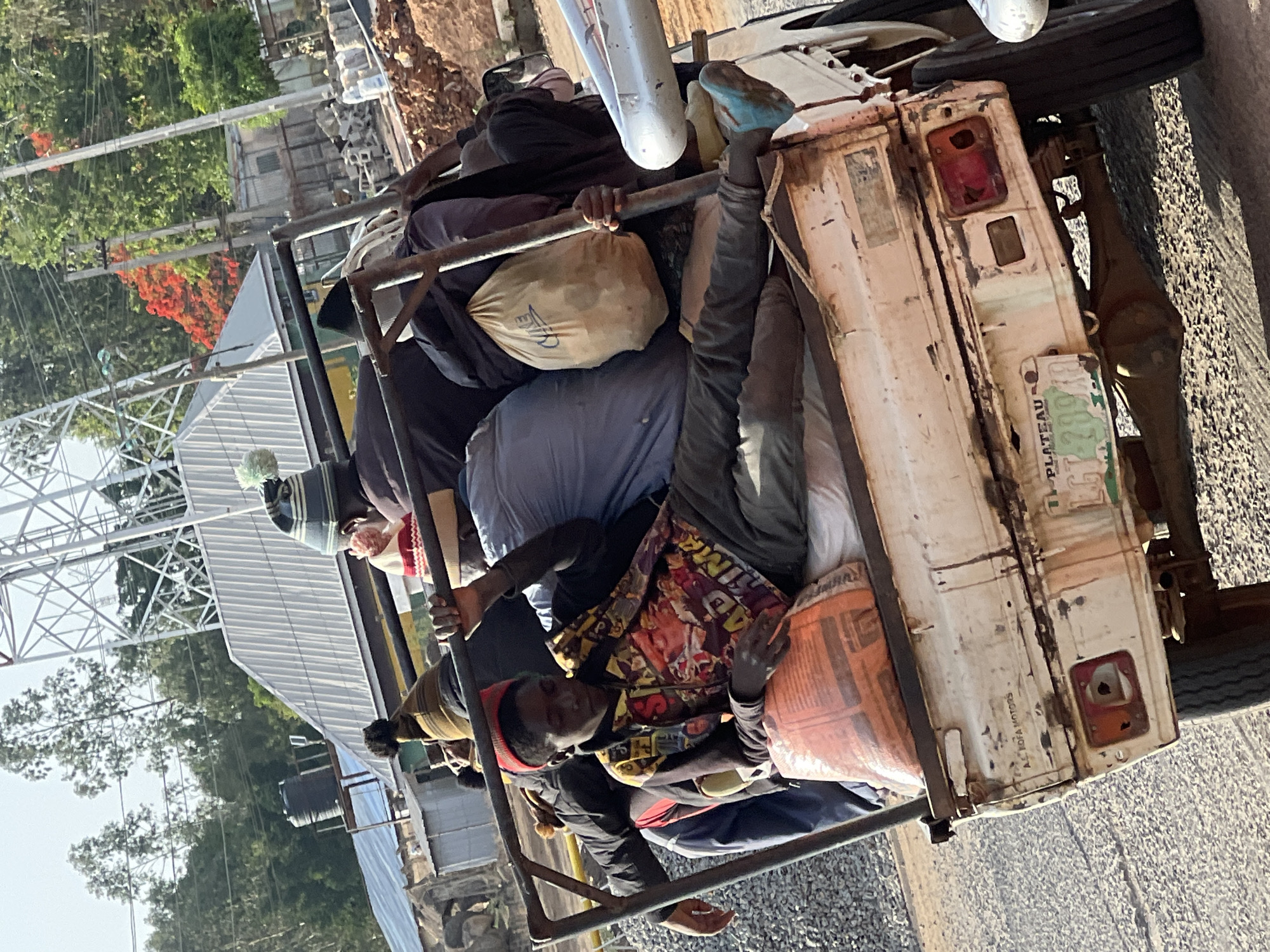 The image depicts a white flatbed truck traveling on a paved road in Jos, Nigeria. The truck bed is overloaded with people and various items, including bags and bundles. Multiple individuals are visible, tightly packed in the truck bed, some holding onto the metal frame surrounding it. The license plate reads "PLATEAU GT-28948". In the background, there are trees, buildings, and utility poles. The sky is bright, suggesting it is daytime.