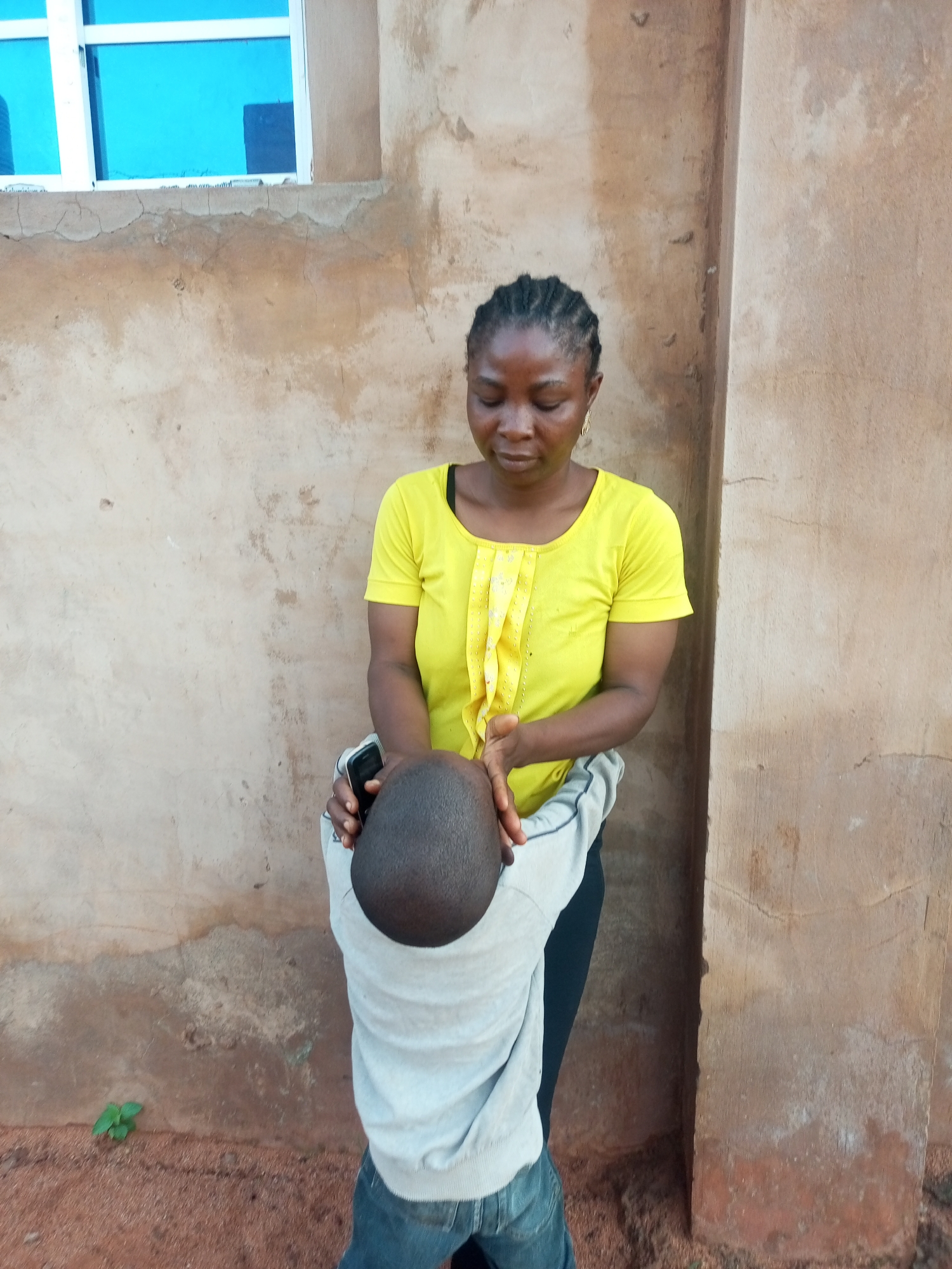 In Tempure, Nigeria, a woman with braided hair and a yellow shirt is standing and leaning over a person who is bent forward. The woman's left hand is resting on the bent-over person's head, while her right hand is holding a black mobile phone. The background consists of a rough, textured wall with a window featuring blue panes above it. The ground is covered in reddish-brown soil.
