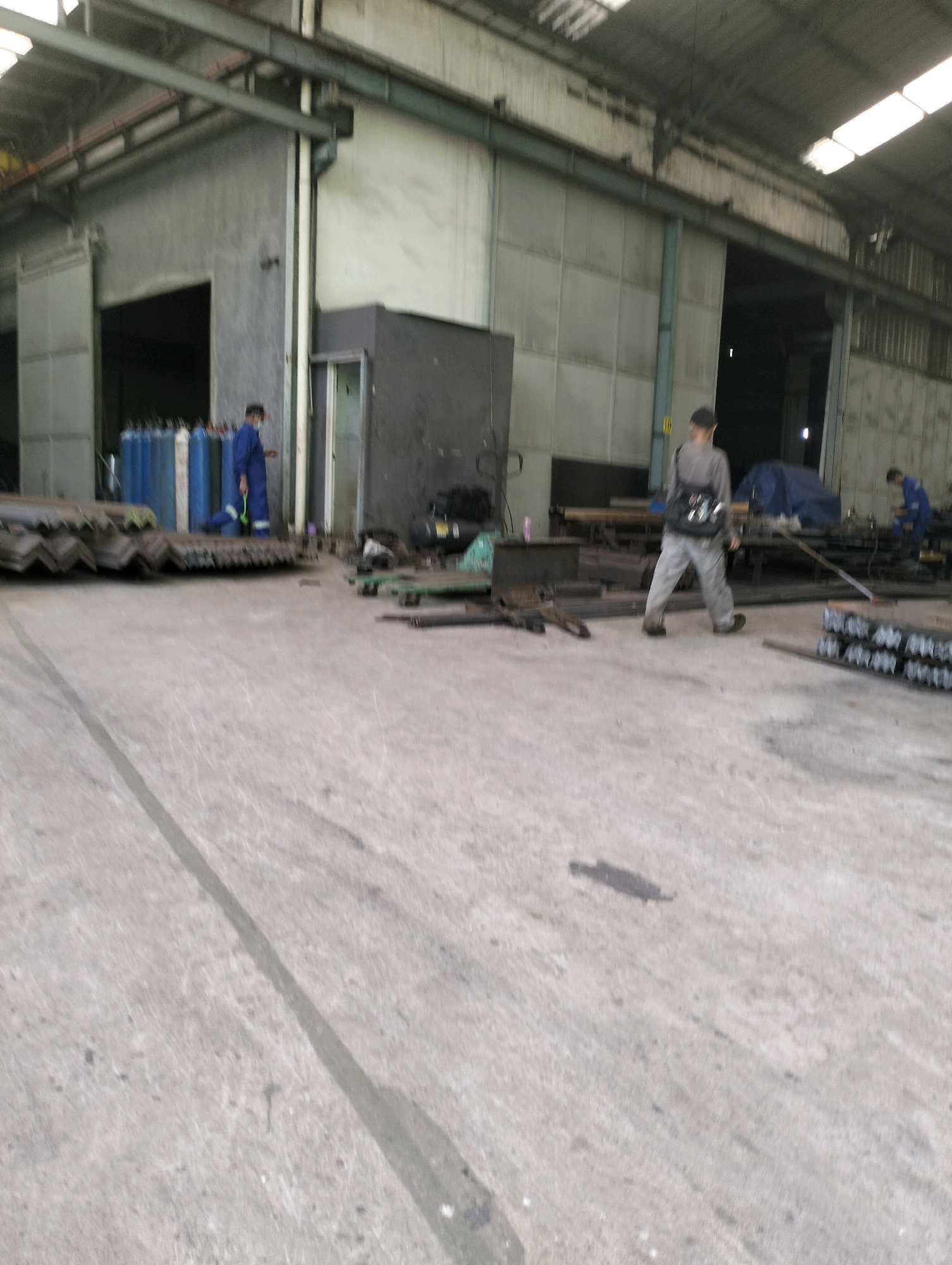 The image shows an interior view of a large industrial building, likely a factory or warehouse, located in Gunung Sindur, Indonesia. The environment is characterized by a concrete floor and high ceilings with exposed beams and translucent roofing panels.
In the left foreground, stacks of dark, angular metal beams are visible. To the left of these beams, a group of tall, blue and white gas cylinders are arranged. A person wearing a blue uniform and a face mask stands near the gas cylinders, facing away from the camera.
In the mid-ground, a person wearing a grey shirt and light-colored pants is walking away from the camera, carrying a dark bag slung over their shoulder. Further in the background, another person in a blue uniform is standing and appears to be working near some machinery. Various pieces of industrial equipment, metal parts, and tarpaulin-covered objects are scattered throughout the space. The overall impression is one of an active industrial setting with materials and personnel involved in manufacturing or storage operations.
