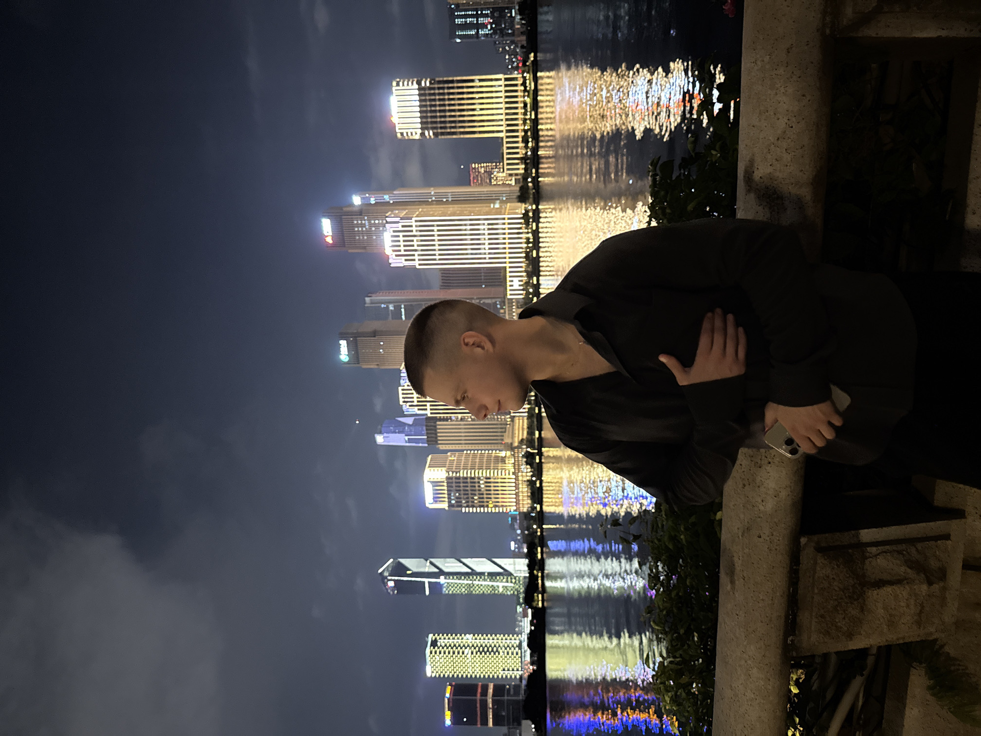The image depicts a nighttime scene in Haizhu District, China, featuring a male individual in the foreground observing a brightly illuminated city skyline across a body of water, likely a river.
In the immediate foreground, a concrete barrier or wall runs horizontally, topped with some dark green foliage. Behind this, the dark water reflects the city lights, creating wavy patterns of yellow, blue, and white illumination.
A male subject is positioned slightly to the right of the frame, facing towards the right and looking at the distant cityscape. He has short, light-colored hair and is wearing a dark, long-sleeved collared shirt with the top buttons unfastened. A thin gold-colored chain or necklace is visible around his neck. His right hand is placed across his chest, and his left hand holds a light-colored smartphone, positioned horizontally near his waist. He is leaning against or standing adjacent to the concrete barrier.
In the midground and background, a modern urban skyline is prominently displayed across the water. Numerous high-rise buildings are visible, extensively illuminated with various lights. Dominant lighting colors are yellow and gold, but white, blue, and red lights are also present on different structures. Several buildings feature distinct lighting patterns, including vertical strips, grids, and illuminated corporate logos (e.g., a red "CRE" logo on one building). The sky above the city is dark with visible cloud formations. The overall scene is a vibrant nighttime view of a metropolitan area.