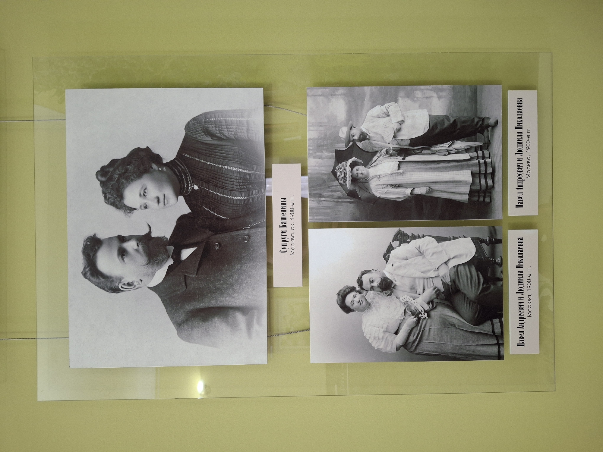 Three vintage black and white photographs are displayed under a clear protective sheet on a light green wall, likely part of an exhibition in Sarapul, Russia.
The leftmost photograph shows a formal portrait of a couple. The woman, positioned at the top right, has dark, curly hair and wears a patterned, possibly ribbed top with decorative elements. She looks directly towards the viewer. The man, at the bottom left, sports a prominent mustache and beard, looking upwards. Associated text identifies them as "Супруги Башенины" (Spouses Bashenins), taken in "Москва, ок. 1900-е гг." (Moscow, approx. 1900s).
The top right photo features a group of people, possibly a family. A man in a hat and jacket gazes down at a seated woman who holds a child. The woman wears a decorated hat, possibly with flowers, and an umbrella or parasol is visible behind them. The accompanying text labels them as "Павел Андреевич и Людмила Николаевна" (Pavel Andreevich and Lyudmila Nikolaevna), from "Москва, 1900-е гг." (Moscow, 1900s).
The bottom right photograph portrays another couple, again identified as "Павел Андреевич и Людмила Николаевна" from "Москва, 1900-е гг.". The man is seated, looking towards the woman, who leans against him, facing the viewer. She is dressed in a light-colored, possibly ruffled or lace garment, while the man wears a light suit jacket.
All photographs capture formal portraits from the early 20th century, displaying the attire and photographic style of that era.