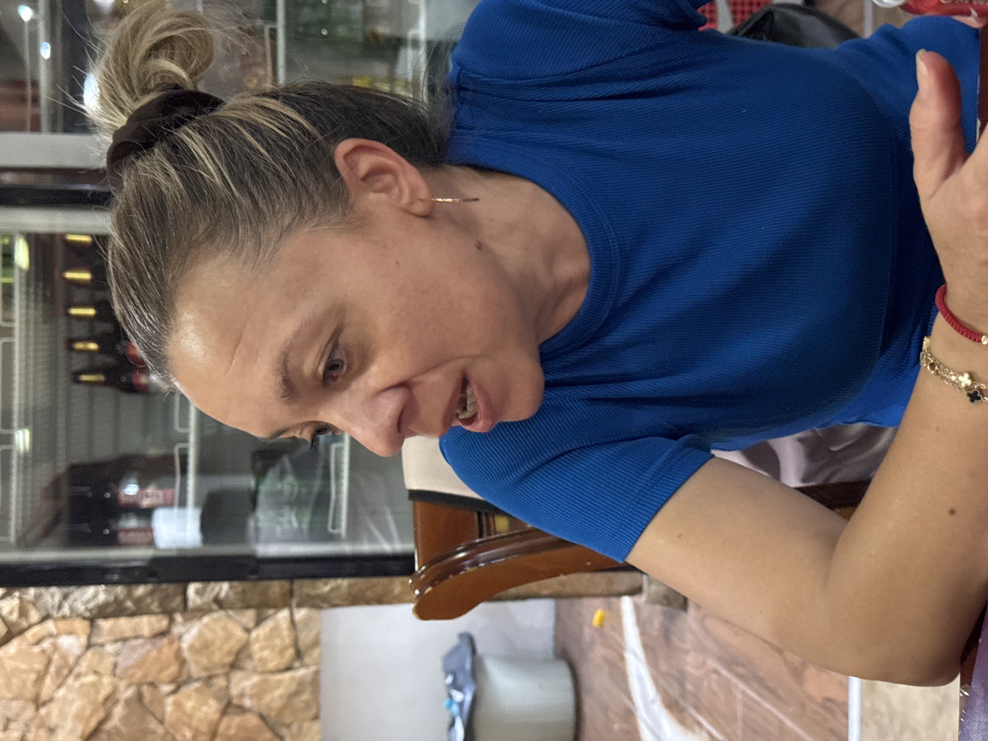 In Sredets, Bulgaria, a medium shot captures a Caucasian woman in her late 30s or early 40s, seated indoors. She has fair skin, light brown hair pulled into a messy bun with some strands loose, and light blue eyes. She is wearing a bright blue, ribbed short-sleeved shirt and a thin gold hoop earring in her left ear. A delicate bracelet with red string and gold charms adorns her right wrist. She is looking to her left, with her mouth slightly open as if speaking.
The background reveals a rustic stone wall on the left and a glass-fronted refrigerator filled with bottles on the right. A portion of a wooden chair with a light-colored cushion is visible in the foreground. The lighting is moderate, with some shadows suggesting an indoor environment. The overall scene appears to be a casual setting, possibly a restaurant or a home.