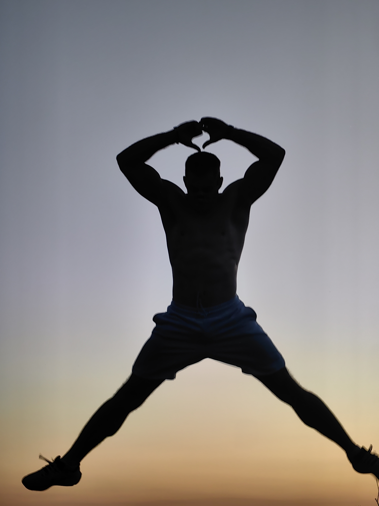 A silhouette of a shirtless man with muscular arms and a lean torso is captured mid-jump against a gradient sky, transitioning from a pale grey at the top to a warm orange-yellow at the bottom, suggesting either dawn or dusk. The man is wearing light blue athletic shorts and dark sneakers. His arms are raised above his head, with his hands forming a heart shape. His legs are spread wide in a dynamic pose, emphasizing the motion of the jump. The background is devoid of any discernible city or specific landmarks, making it impossible to determine a location within Varna, Bulgaria, or any other city. The focus is solely on the silhouette and the man's athletic action.