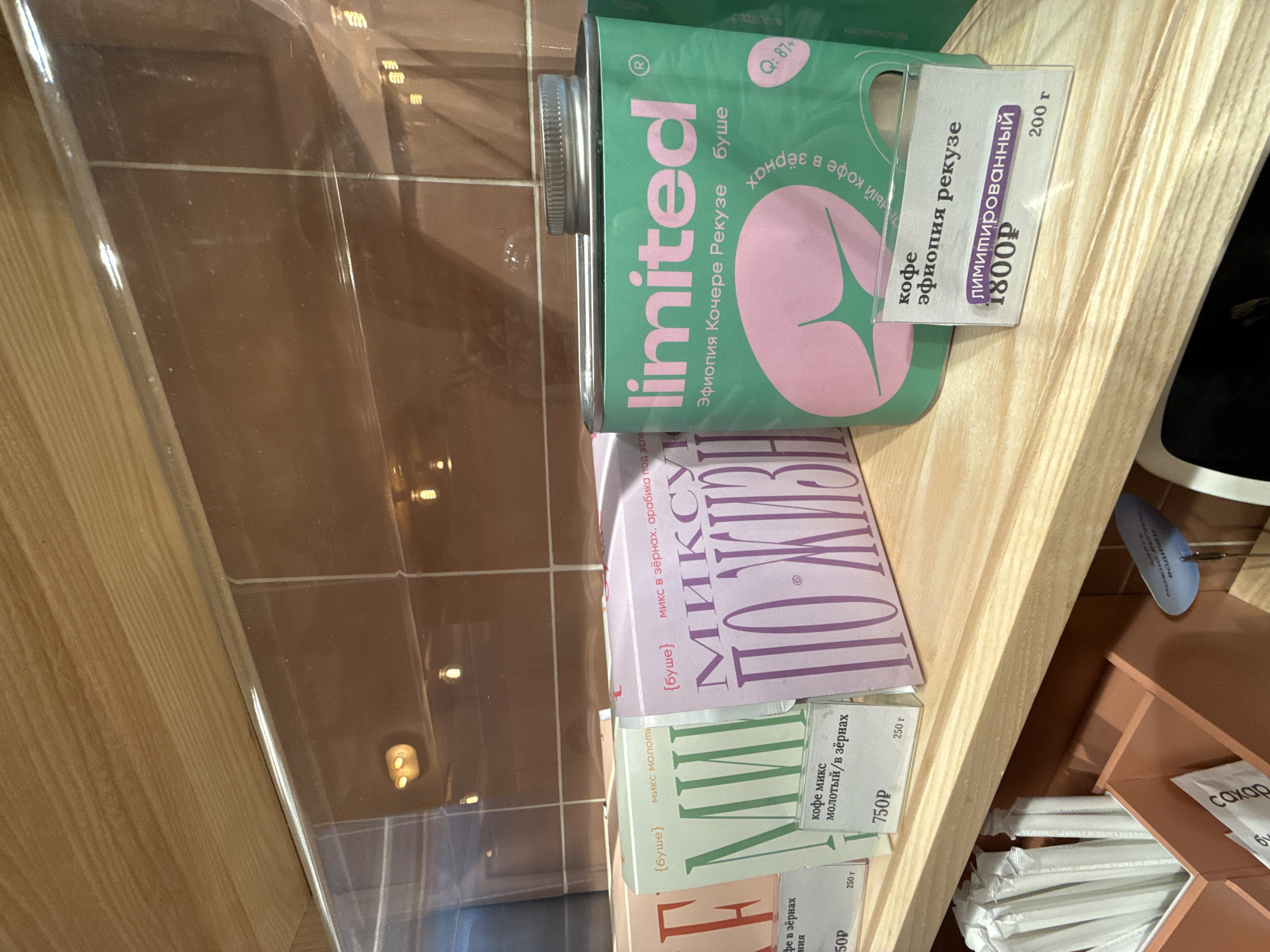 Here is a forensic description of the image:
The image depicts a selection of coffee products in Saint Petersburg, Russia. A green, cylindrical "limited" brand coffee container with a pink logo, labeled "Эфиопия Кочере Рекузе" (Ethiopia Kochere Rekuze) and marked with a price tag of 1800P for 200g, is prominent. Below it are boxes of coffee mix, labeled "МИКСУ ПОЖИВИ", and "МИИ", labeled "кофе микс молотый/в зёрнах" (coffee mix ground/in grains) and priced at 750P for 250g. A white sugar packet, drinking straws, and other coffee-related supplies are also visible. The setting appears to be a store shelf or counter, with a brown tiled wall in the background.