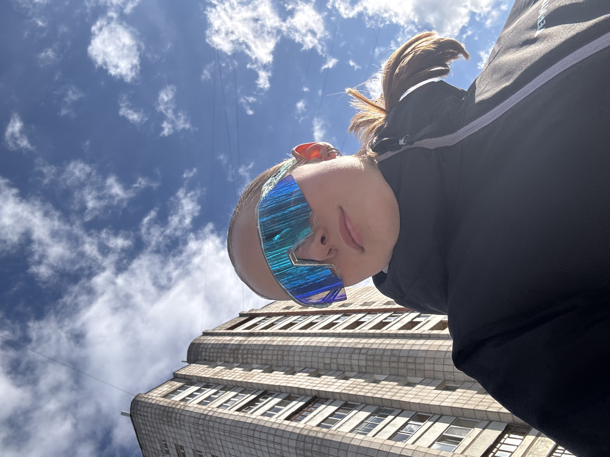 A person is captured in a low-angle shot, looking down towards the camera, against a bright blue sky with scattered white clouds, in Chaikovsky, Russia. The individual is primarily visible from the head to the upper torso, wearing large, iridescent blue, futuristic-style sunglasses that obscure their eyes and upper face. Their light brown hair is pulled back, and their lips are slightly parted, suggesting a relaxed expression. They are dressed in dark-colored athletic clothing, possibly a jacket, featuring a gray or white stripe and zipper detail on the shoulder. Faint, light-colored text, potentially a brand name (e.g., "RUNNER"), is partially visible on the shoulder.
In the background, a large, multi-story building with a textured, tiled facade and numerous rectangular windows rises upwards, occupying the lower right and center of the frame. The building's architectural style features stepped-back upper floors, hinting at an older design. Utility wires or power lines are visible stretching across the clear sky. The scene is illuminated by bright daylight, indicating a sunny day. The individual appears to be posing for a photograph in an urban or semi-urban outdoor setting.