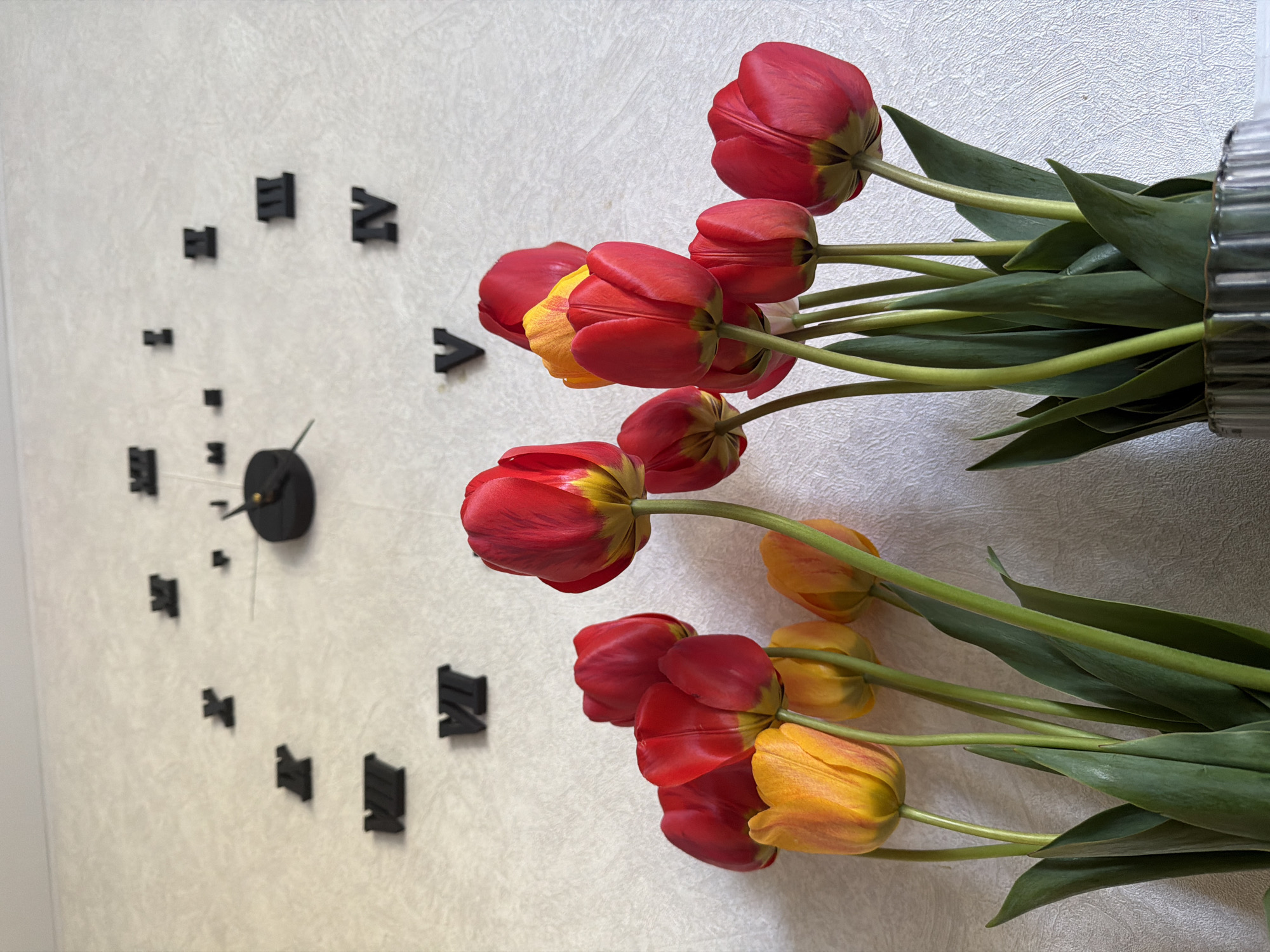 A still life photograph depicts a wall clock next to a bouquet of tulips. The clock is a modern design with black Roman numerals adhered to the wall. The bouquet features red and yellow tulips with green stems and leaves. The flowers are arranged in a vase which is out of view, only a small part is seen on the right. The location is indeterminate, and there is no evidence in the image to indicate it is Tokkari, Russia. There are no people visible.