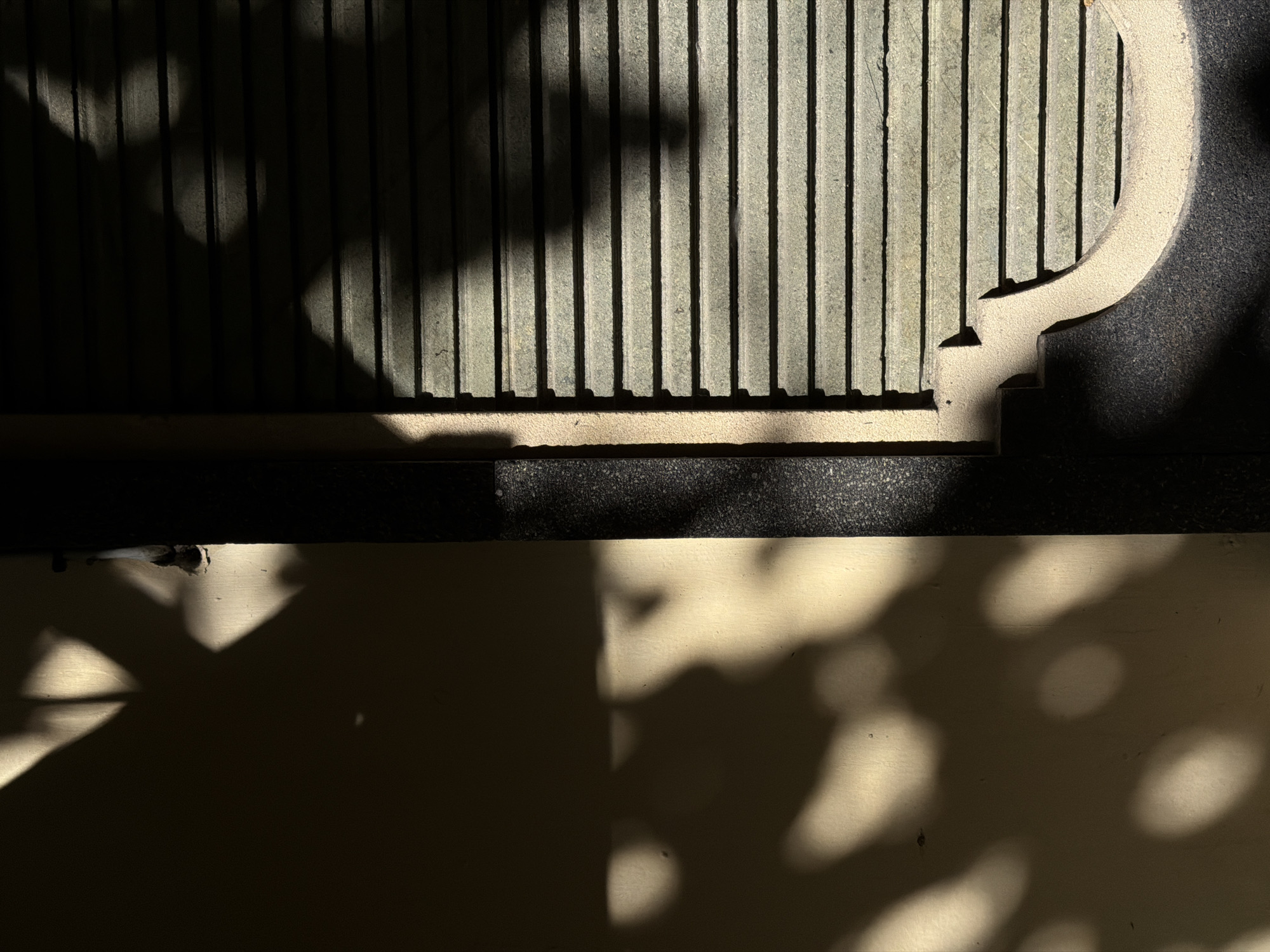 The image shows a close-up of a concrete structure, likely outdoors, in North Kuta, Indonesia. The structure features vertical, ribbed concrete panels with some sort of curved edge ornamentation. Shadows from foliage partially obscure the panels. Below the concrete structure, a flat, light-colored surface is visible, also with shadows.