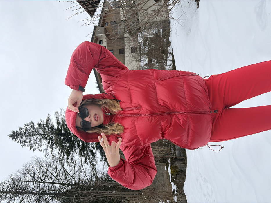 A blonde-haired individual, wearing a red hooded puffer jacket and matching red pants, stands in a snow-covered landscape located in La Clusaz, France. The subject wears black square-frame sunglasses and protrudes their tongue slightly. The left hand is raised near the temple, while the right hand forms a "rock on" gesture, holding a lit cigarette or cigarillo between the index and middle fingers. The background features several bare deciduous trees and evergreen conifers on the left, and multi-story wooden chalet-style buildings with balconies on the right, interspersed with leafless bushes. The ground is uniformly covered in snow under an overcast sky.