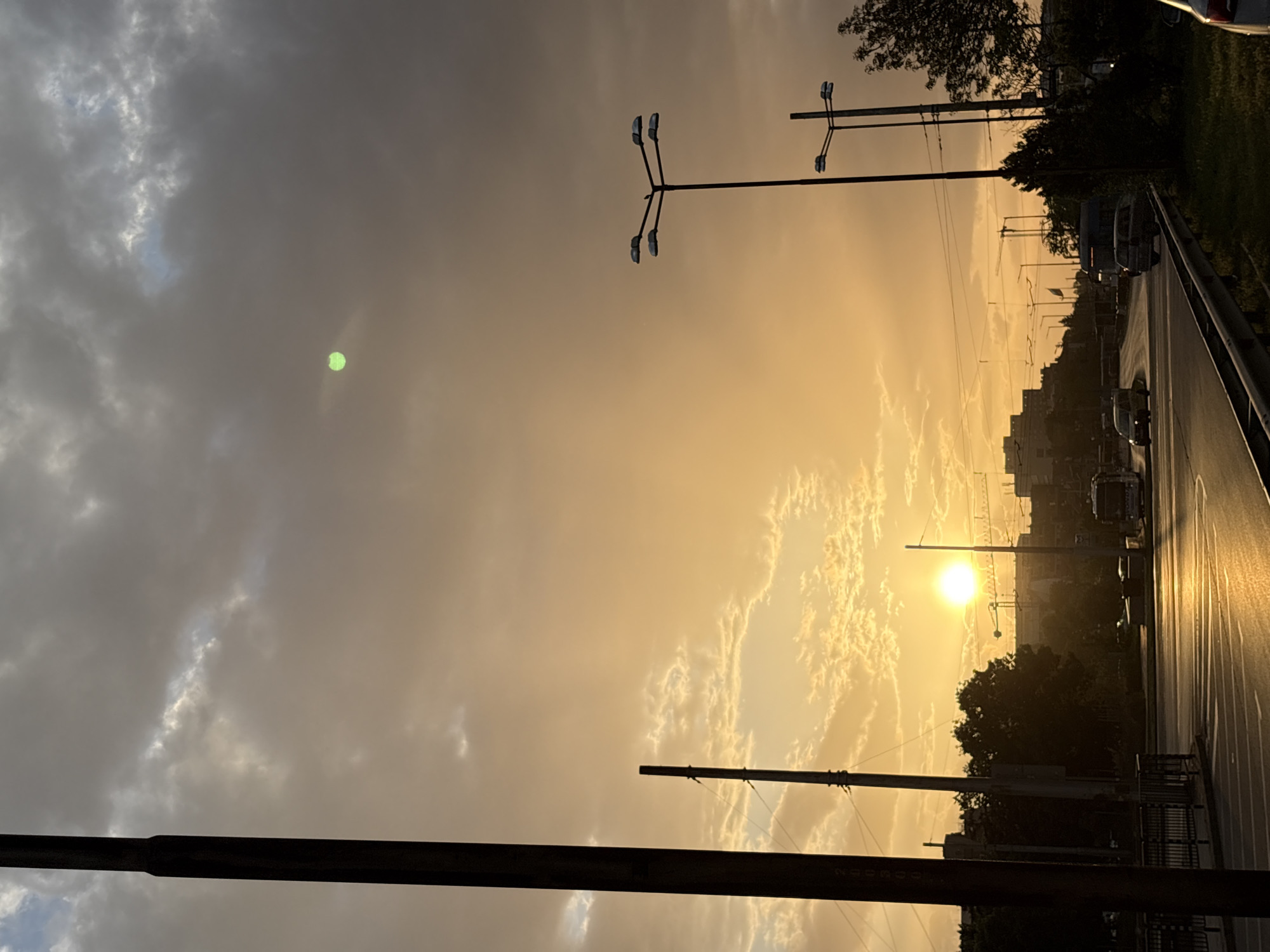 The image depicts a street in Sofia, Bulgaria at sunset. The sky is filled with clouds, with the sun appearing as a bright orb near the horizon. The light casts a golden glow over the scene. Streetlights are visible along the road. Vehicles are present on the road, suggesting traffic flow. Buildings are visible in the background. A green reflection is visible in the top left corner. The photo is taken from a low angle, possibly street level, with what appears to be a pole or rail obscuring the bottom portion of the frame. The light reflecting off of a surface on the bottom right appears to be reflective and wet.