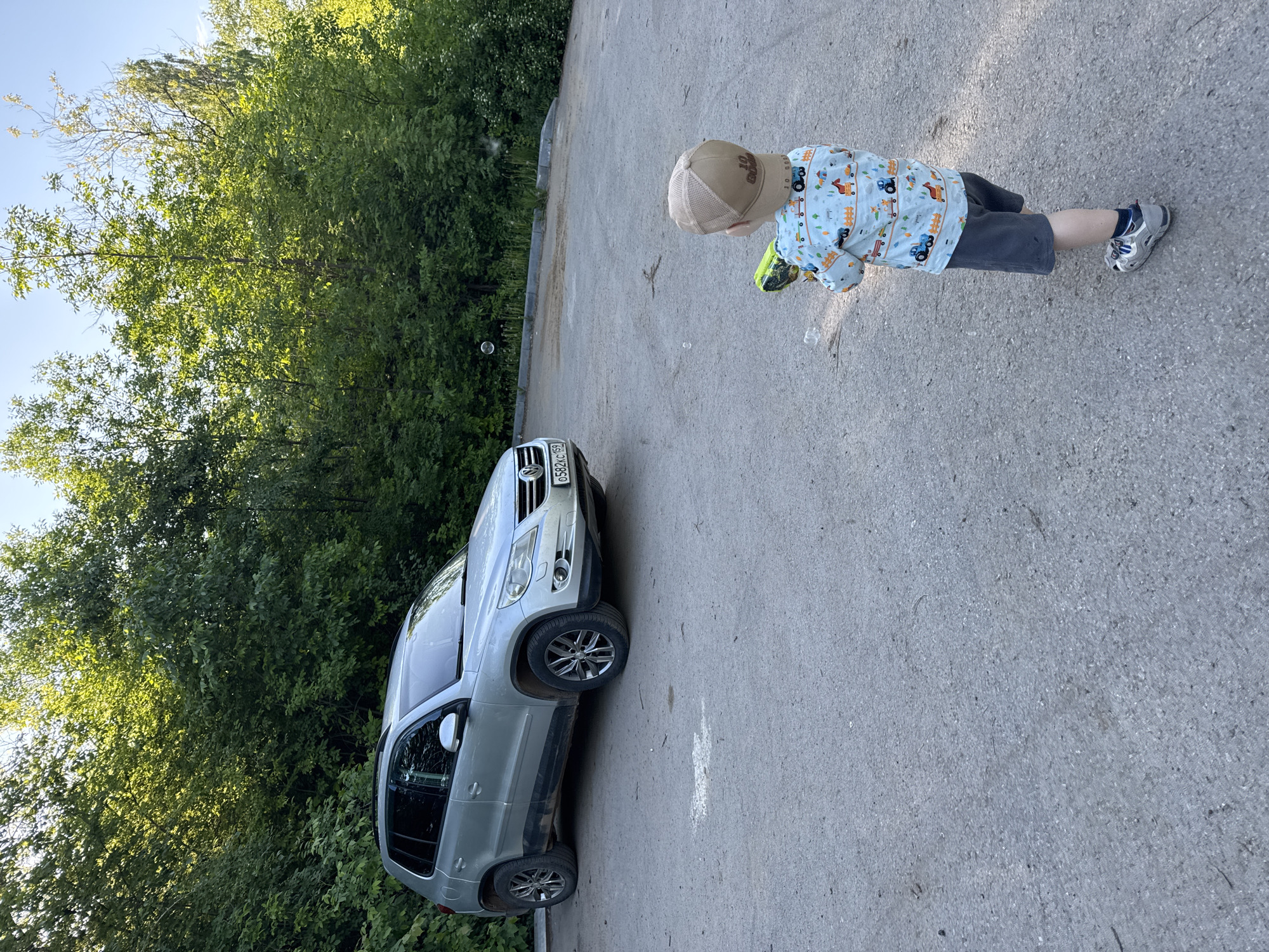 The image captures an outdoor scene in the city of Vereshchagino, Russia, featuring a young child and a parked silver Volkswagen Tiguan.
A child, viewed from behind, stands in the foreground on a grey asphalt surface, holding a green and black bubble gun. The child wears a light blue long-sleeved shirt patterned with various cartoon vehicles and trees, dark grey shorts, and light grey athletic shoes with blue accents. A tan mesh baseball cap is worn on the child's head, displaying text that is partially obscured but appears to include "10 GOALS". Several spherical bubbles are visible floating in the air, originating from the bubble gun.
On the left side of the frame, a silver Volkswagen Tiguan SUV is parked on the same asphalt surface. The vehicle exhibits dirt and dust on its lower side panels. A Russian license plate, "O582XC 159," is mounted on the front bumper.
The background is dominated by a dense wall of vibrant green trees and bushes, indicating a wooded area or an overgrown natural embankment. Sunlight brightly illuminates the upper foliage, suggesting a clear, sunny day. The asphalt ground has a textured appearance with some scattered debris and lighter, faded patches.