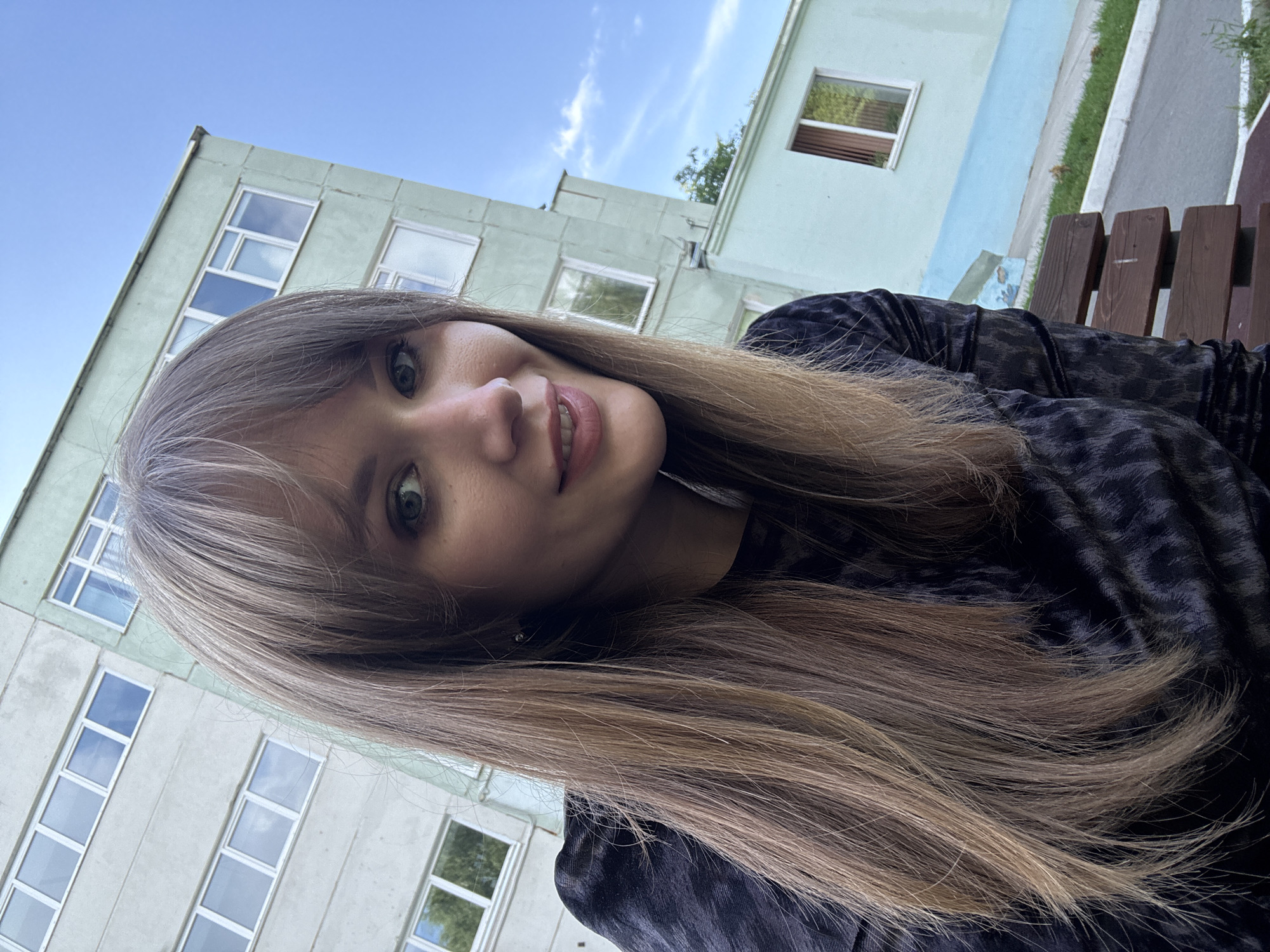 A medium shot shows a young Caucasian woman with long, light brown hair and bangs, smiling at the camera. She is wearing a black patterned top. In the background, there is a light green apartment building with multiple windows and a bright blue sky with a few white clouds. To the right, there is a lower section of the building painted light blue, and a wooden bench with green grass and a sidewalk in front of it. The scene is set on a street in Chaikovsky, Russia.