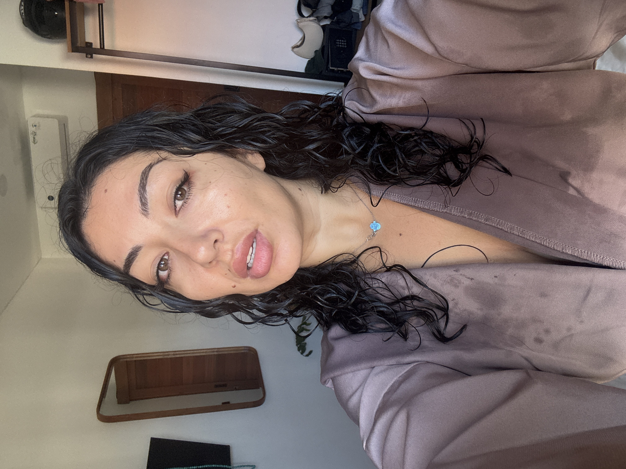 A woman with wet, dark, curly hair is taking a selfie in what appears to be a hotel room in Kuta Selatan, Indonesia. She is wearing a mauve-colored robe that is damp in places. She has on a silver necklace with a light blue pendant. In the background, there is a mirror with a wooden frame, a wall-mounted air conditioner, and clothing draped over a rack.