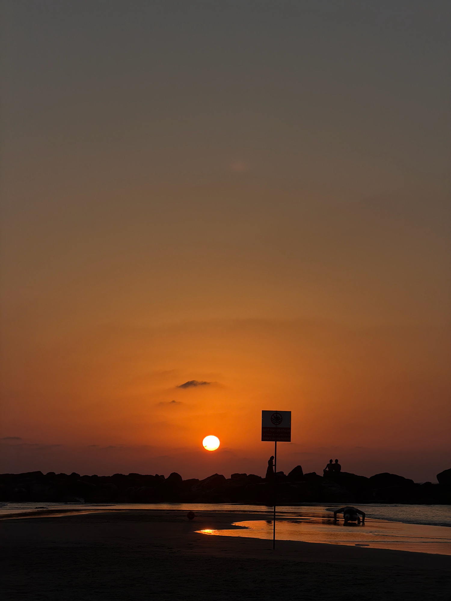 A vertical, low-angle shot captures the vibrant orange and gray hues of a sunset over the Mediterranean Sea at Tel Aviv's Gordon Beach. The sun, a bright orb, hovers low on the horizon, casting a warm glow that reflects on the wet sand. Silhouetted against the colorful sky are several figures: a person standing near a sign, two people sitting on rocks, and another person in a low, crawling posture near the water's edge. The dark rocks that line the shore create a stark contrast with the luminous sky and sea. A partially visible red ball lies on the sand. The scene suggests a peaceful evening at the beach, with people enjoying the last moments of daylight.