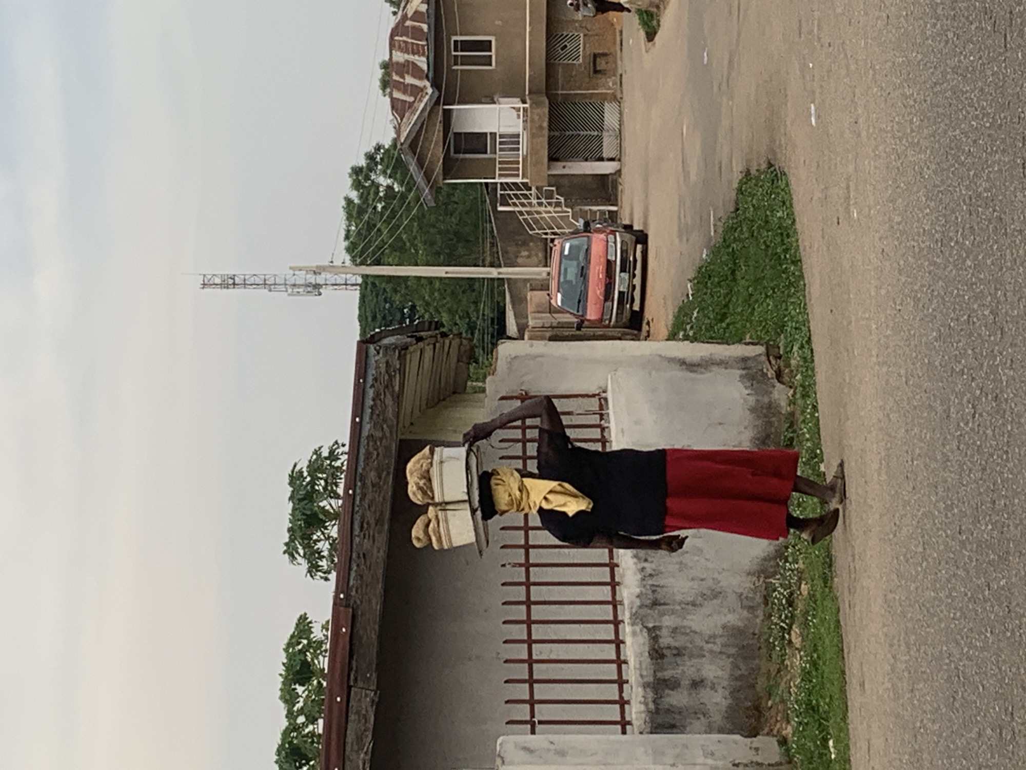 A woman carries a load on her head while walking on a paved road in Gura Top, Nigeria. She is wearing a black top and red skirt with sandals. The load appears to be buckets covered with fabric. She walks past a low wall with a red metal gate. Behind the wall is a building with a rusted roof. Across the road is a multi-story building with exterior stairs, a red car parked out front. In the background, a cell tower is visible. The sky is overcast.