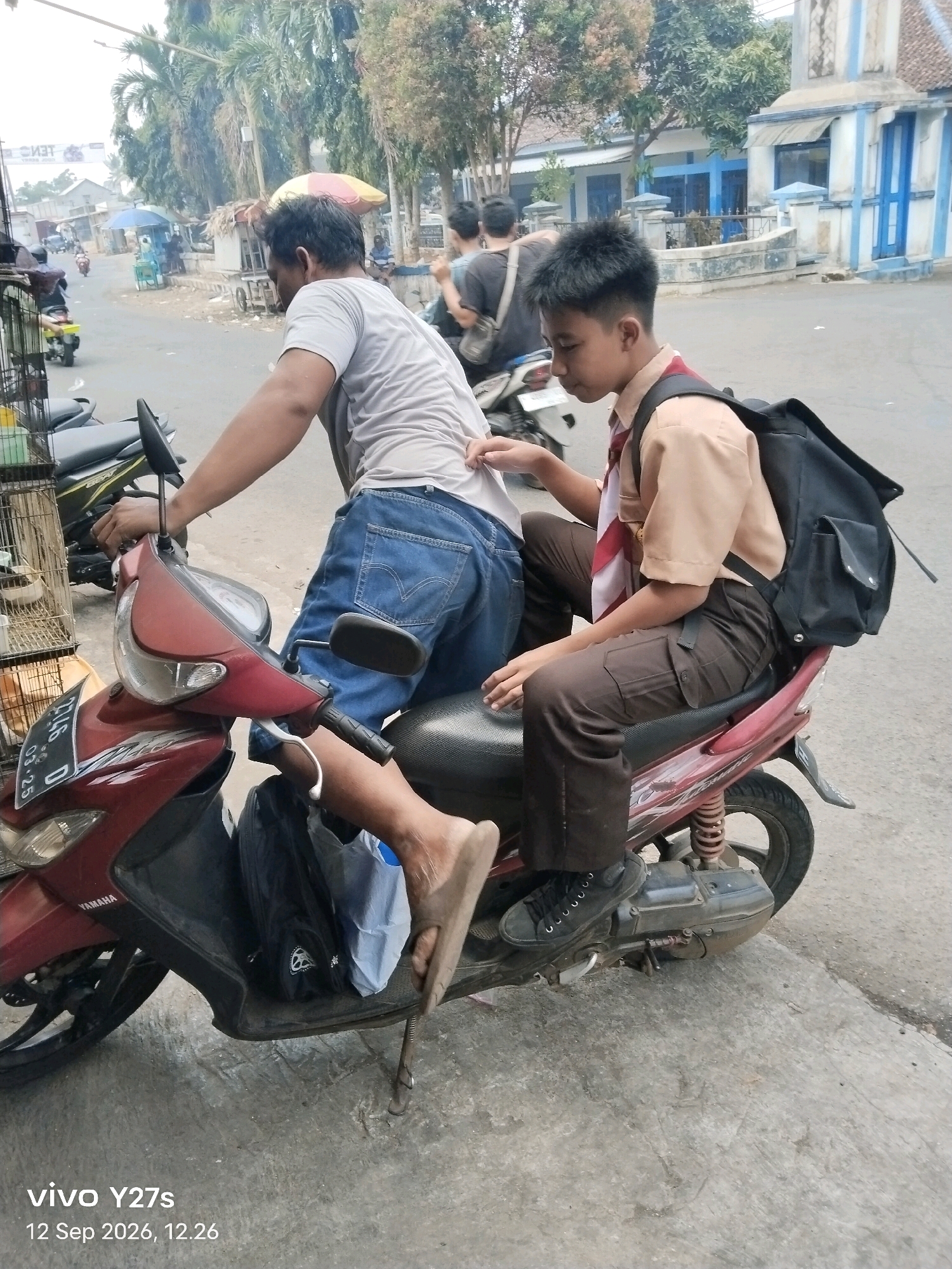 In Trimulyo, Indonesia, two individuals are on a maroon and red scooter. The scooter has a license plate that reads "BG 2746 D". A person wearing a grey t-shirt and blue jeans is seated at the front of the scooter, facing away from the camera, with their right hand on the handlebar and their left hand resting on the back of the seat. They are barefoot. A younger person, appearing to be a student in a tan uniform shirt and brown pants, is seated behind the first person. This student has a red and white scarf around their neck and is wearing a black backpack. Their left hand is resting on the scooter's seat, and their right arm is bent and positioned on their leg. The scooter is parked on a paved surface, possibly a road or sidewalk, with a concrete curb in the foreground. In the background, there are buildings with blue trim, trees, and other scooters. The timestamp "12 Sep 2026, 12.26" and "vivo Y27s" are overlaid on the bottom left of the image.
