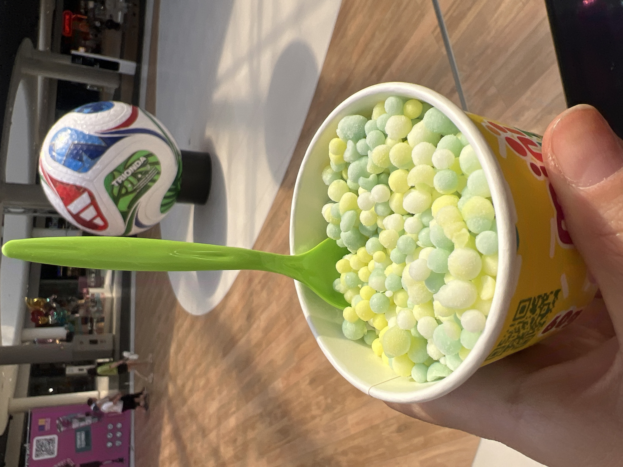 A close-up shot shows a hand holding a cup of bubblegum-flavored ice cream. The ice cream is served in small, round balls and is a pastel mix of green, yellow, and white. A bright green plastic spoon is stuck in the ice cream. In the background, blurred figures of people can be seen walking through a shopping mall. A large soccer ball is displayed on a pedestal, and colorful balloons float near the ceiling. The floor is made of polished wood. The lighting suggests an indoor environment with artificial light. The image is taken from a low angle, looking up at the cup. The overall scene appears to be a casual setting within a commercial area. There is no indication that this location is in Kyiv, Ukraine.