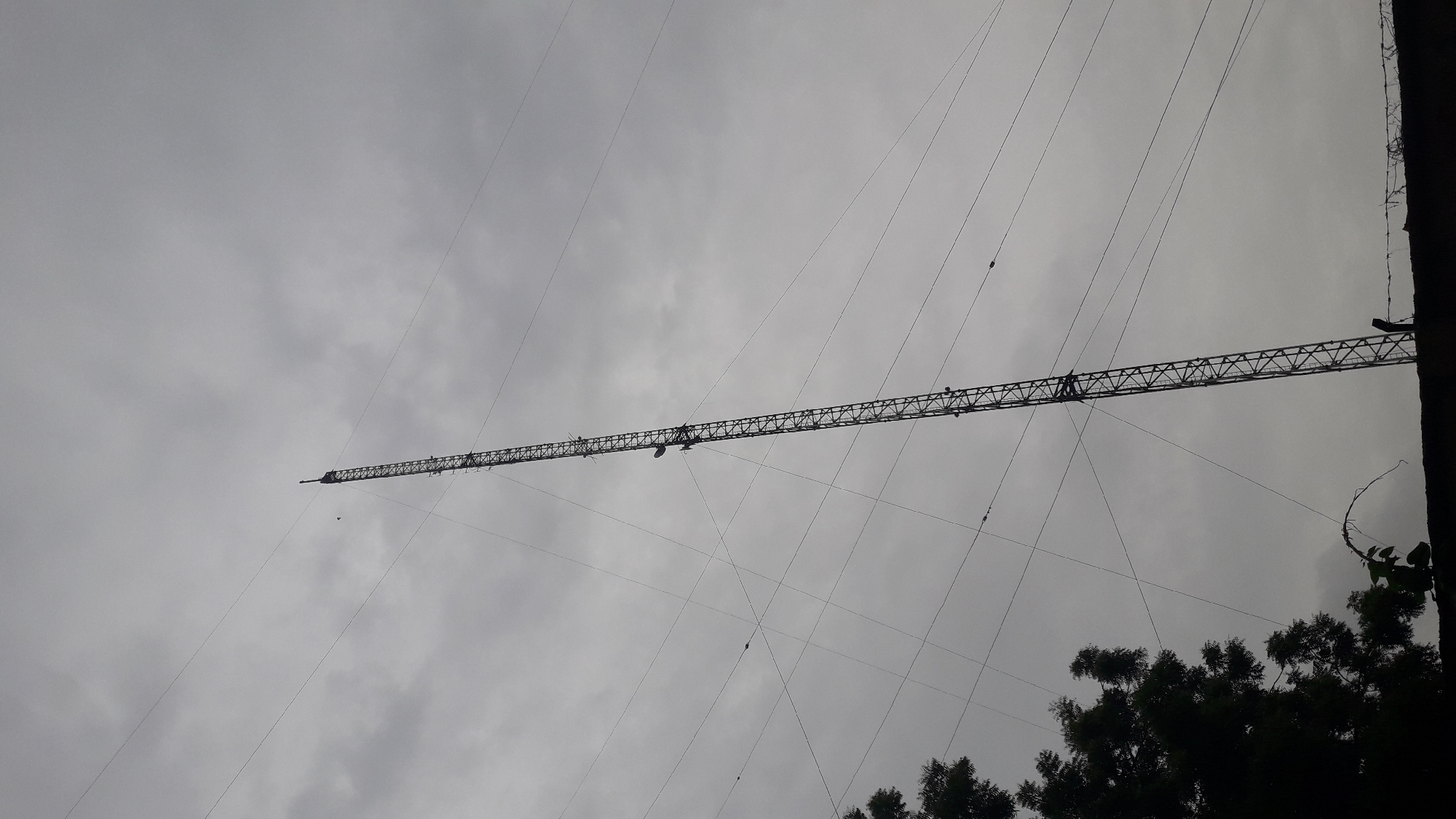 The image shows a long, horizontal lattice boom crane extending across the sky. Thin support cables are strung from the boom down towards the ground. The sky is overcast with grey clouds. Trees and a dark structure, possibly a building, are visible in the lower right corner of the frame. Given the request to include the location of Onitsha, Nigeria, it can be inferred that this is where the photograph was taken.