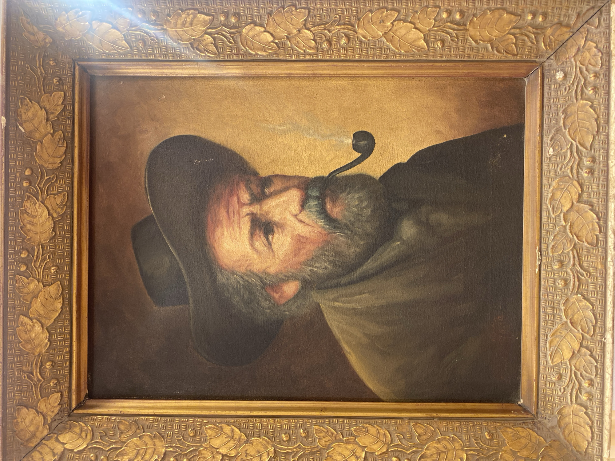 This is a framed oil painting of a man wearing a hat and smoking a pipe. The man has a beard and is looking to the right. The painting is housed in an ornate gold frame with leaf motifs. The background of the painting is a gradient of brown and gold. There is a faint plume of smoke rising from the pipe. The painting is displayed in an indoor setting. There is no indication in the image that this painting is located in the city of Legenda, Egypt.