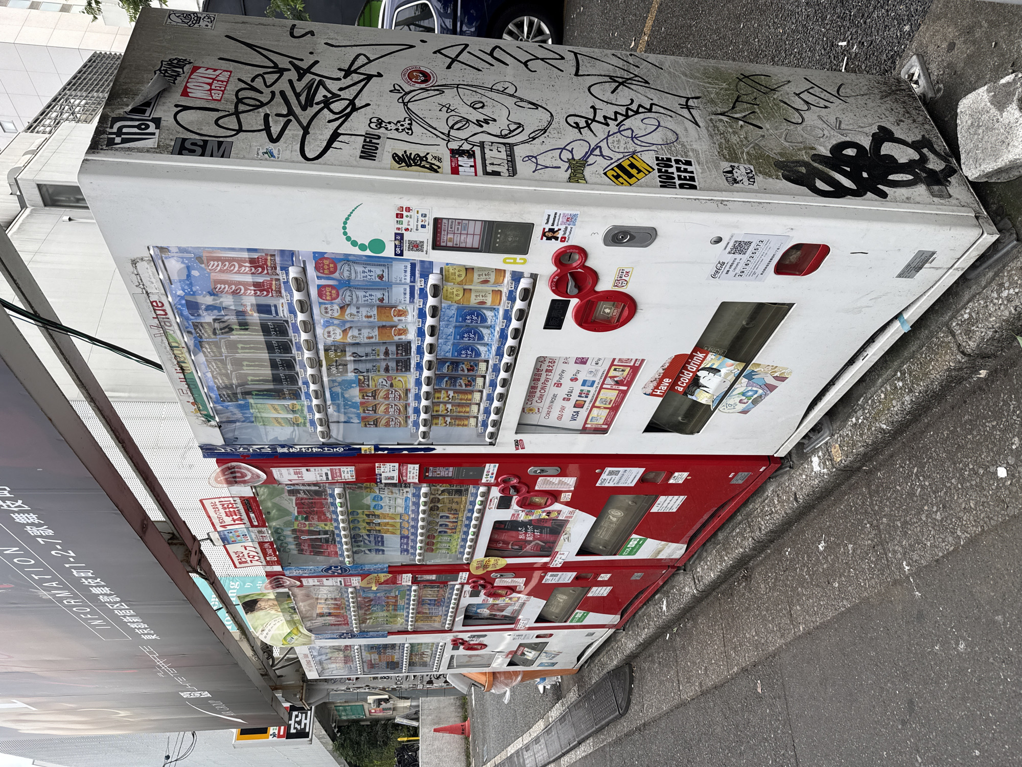 The image depicts a row of vending machines on a city street in Shinjuku, Japan. The vending machines are positioned alongside a sidewalk, with a street visible in the foreground. In the background, a multi-story building can be seen. The vending machines are stocked with various beverages, and some of them are adorned with graffiti. One of the vending machines prominently displays the Coca-Cola logo and payment options, including Paypay, R-Pay, and UnionPay. The street appears to be a commercial area, given the presence of vending machines and the building in the background.
