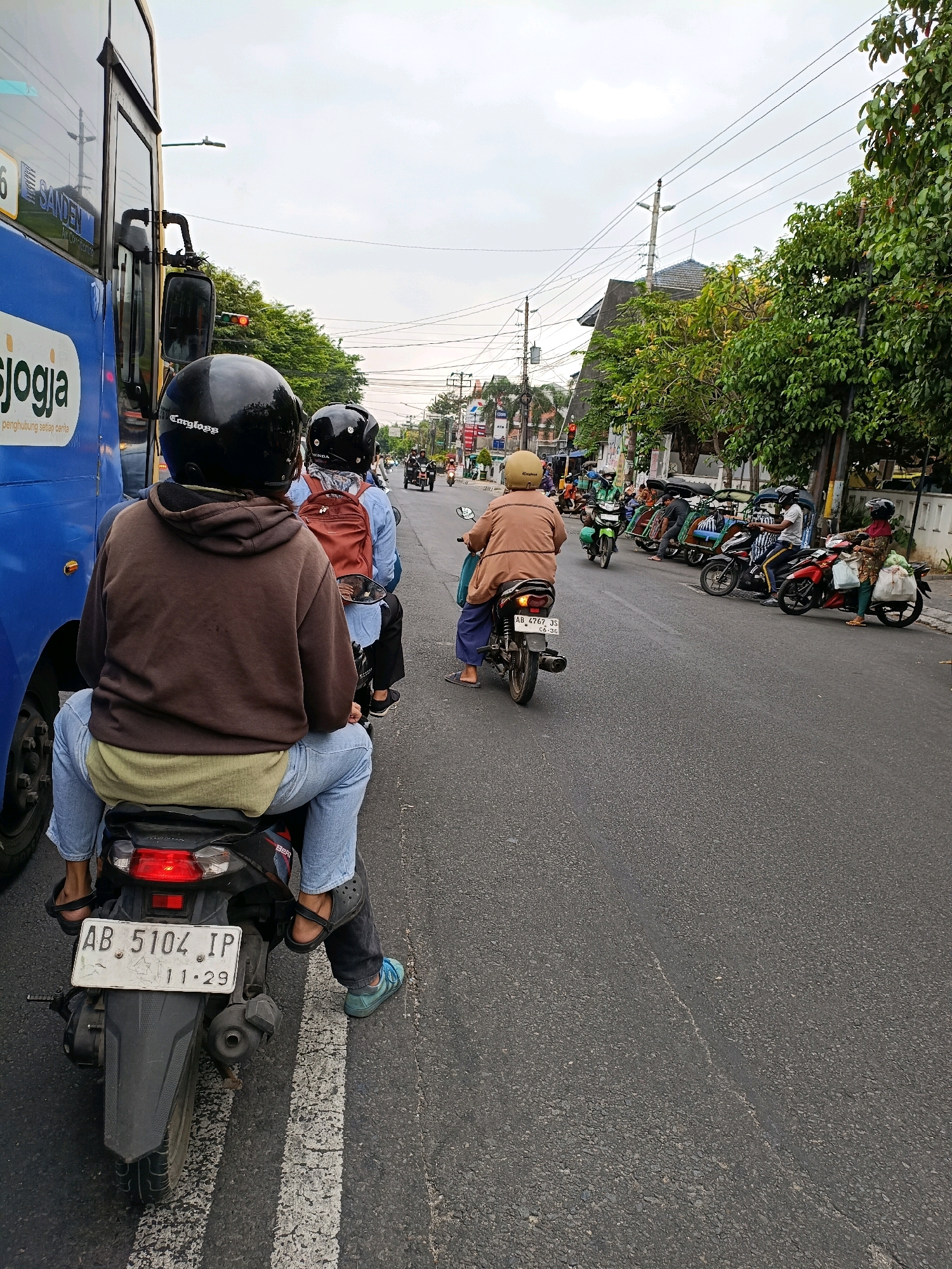 The image captures a street scene in Yogyakarta, Indonesia, under an overcast sky. In the foreground, a motorcycle with two riders is stopped at what appears to be a traffic light. The rider in the back, wearing a brown hooded jacket and blue jeans, is the primary subject of the shot. They are wearing a black helmet. The passenger in front of them is wearing a blue shirt and a brown backpack, also with a black helmet. To the left, a portion of a blue bus with "jogja" logo is visible, indicating its presence in the city. The street itself is paved and marked with white lines. In the background, several other motorcycles, some with passengers, are lined up, suggesting traffic congestion or a waiting period. Buildings and trees line the side of the street, and power lines are visible overhead. A traffic light is also present, likely contributing to the stop.