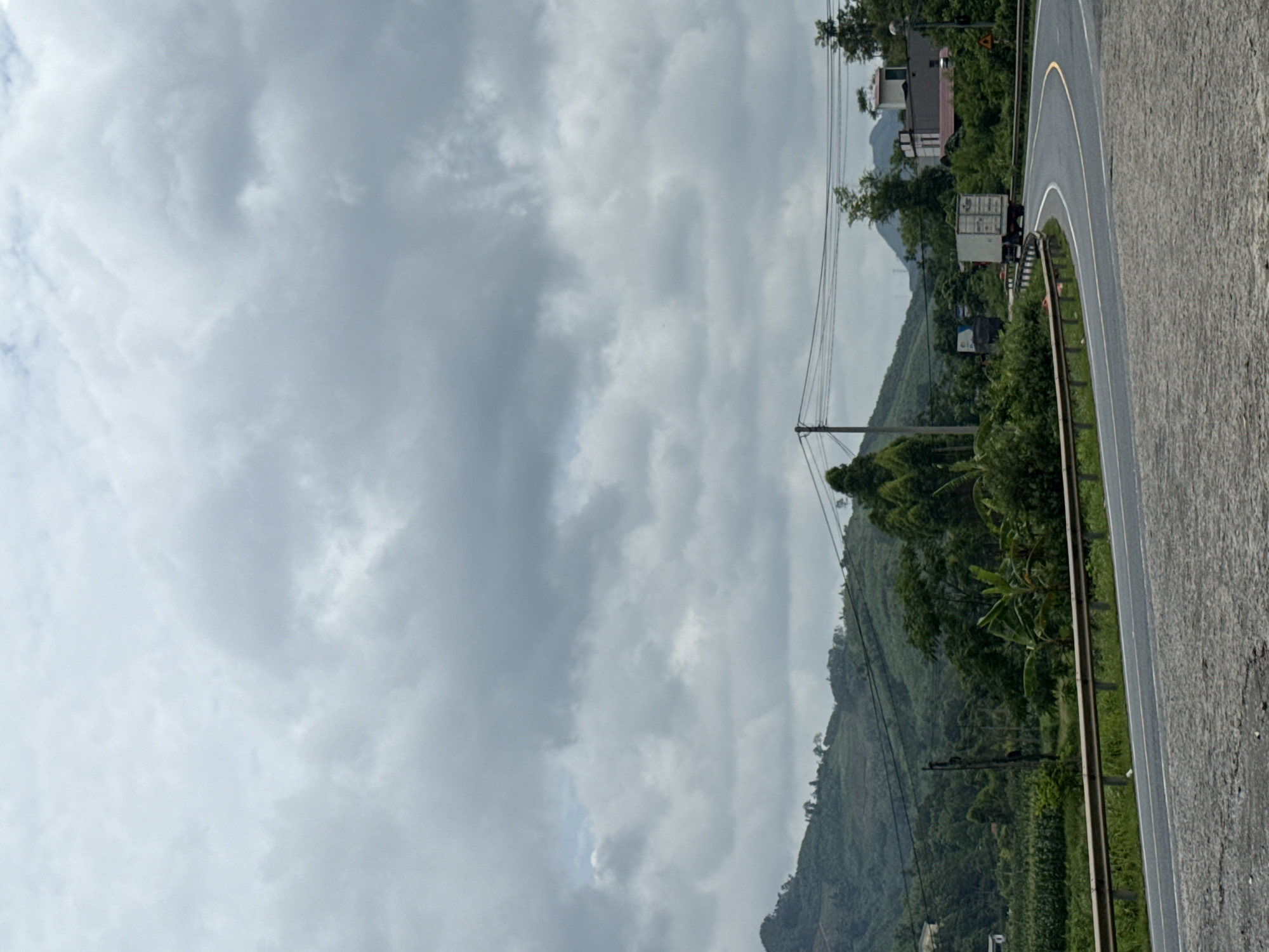 The image shows a roadside scene in Mai Sao, Vietnam. The right side of the image features a grey asphalt road curving slightly. A metal guardrail runs along the road's edge. Beyond the guardrail, the landscape consists of lush green vegetation covering a hillside. Power lines are visible extending across the scene, supported by utility poles. In the background, there are buildings and possibly a mountain obscured by overcast skies. The sky dominates much of the upper left portion of the image and is filled with heavy, grey clouds. A white shipping container can be seen on a truck along the road.