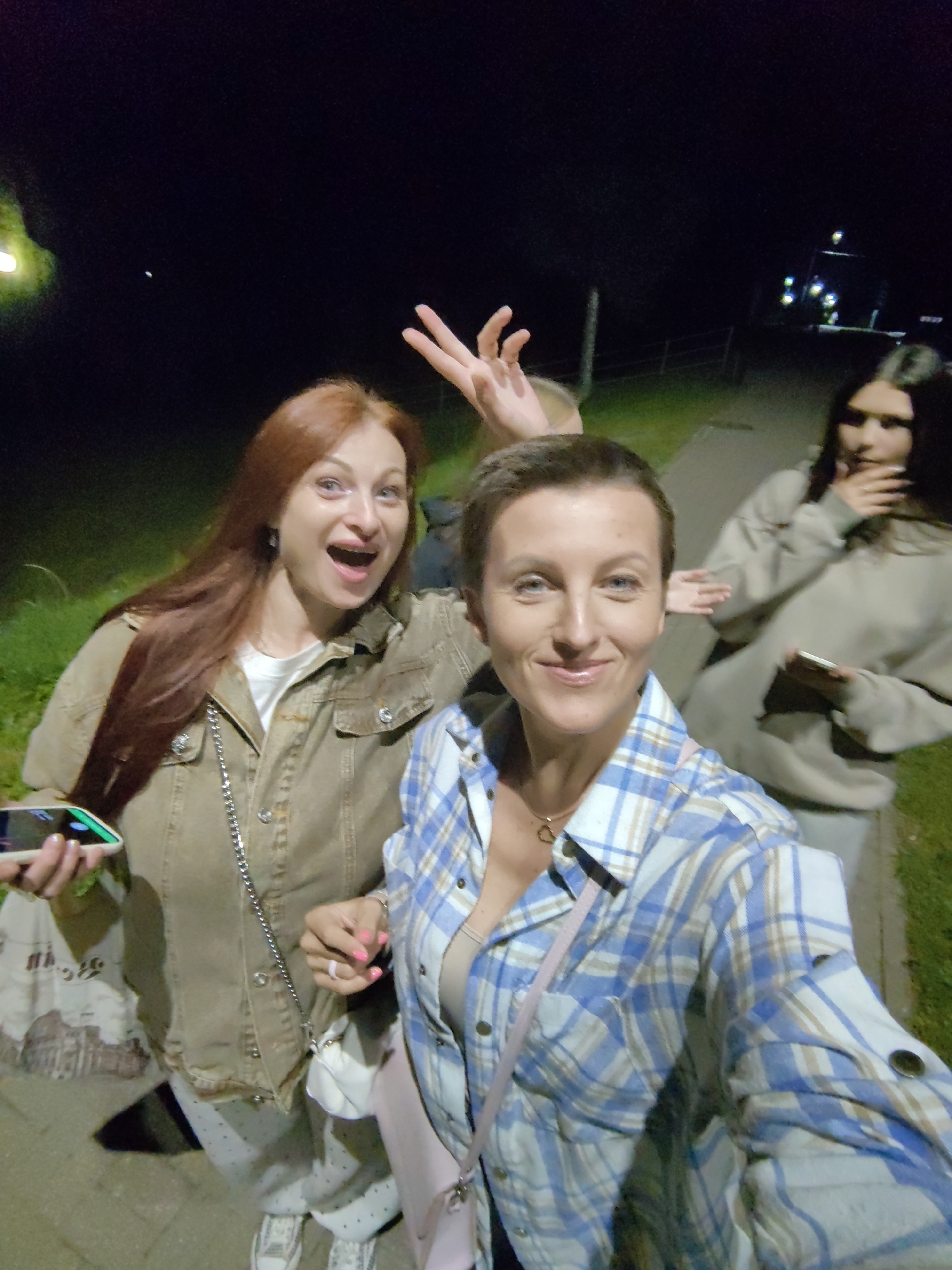Three women are captured in a selfie-style photograph at night, seemingly on a paved pathway bordered by grass. The woman in the foreground, with short, dark hair, is smiling directly at the camera. She is wearing a blue and white plaid shirt over a beige tank top and a pink cross-body bag. To her left, a woman with long, reddish-brown hair is looking upwards with her mouth open in an excited expression, wearing a brown denim jacket over a white t-shirt. Her left hand is raised with fingers spread. Further in the background, a third woman with dark hair is partially visible, with her hand covering her mouth and holding a smartphone. The lighting suggests artificial illumination from streetlights in the distance, with some ambient light contributing to the visibility of the scene. The environment appears to be an outdoor public space in the city of Myadzyel, Belarus.