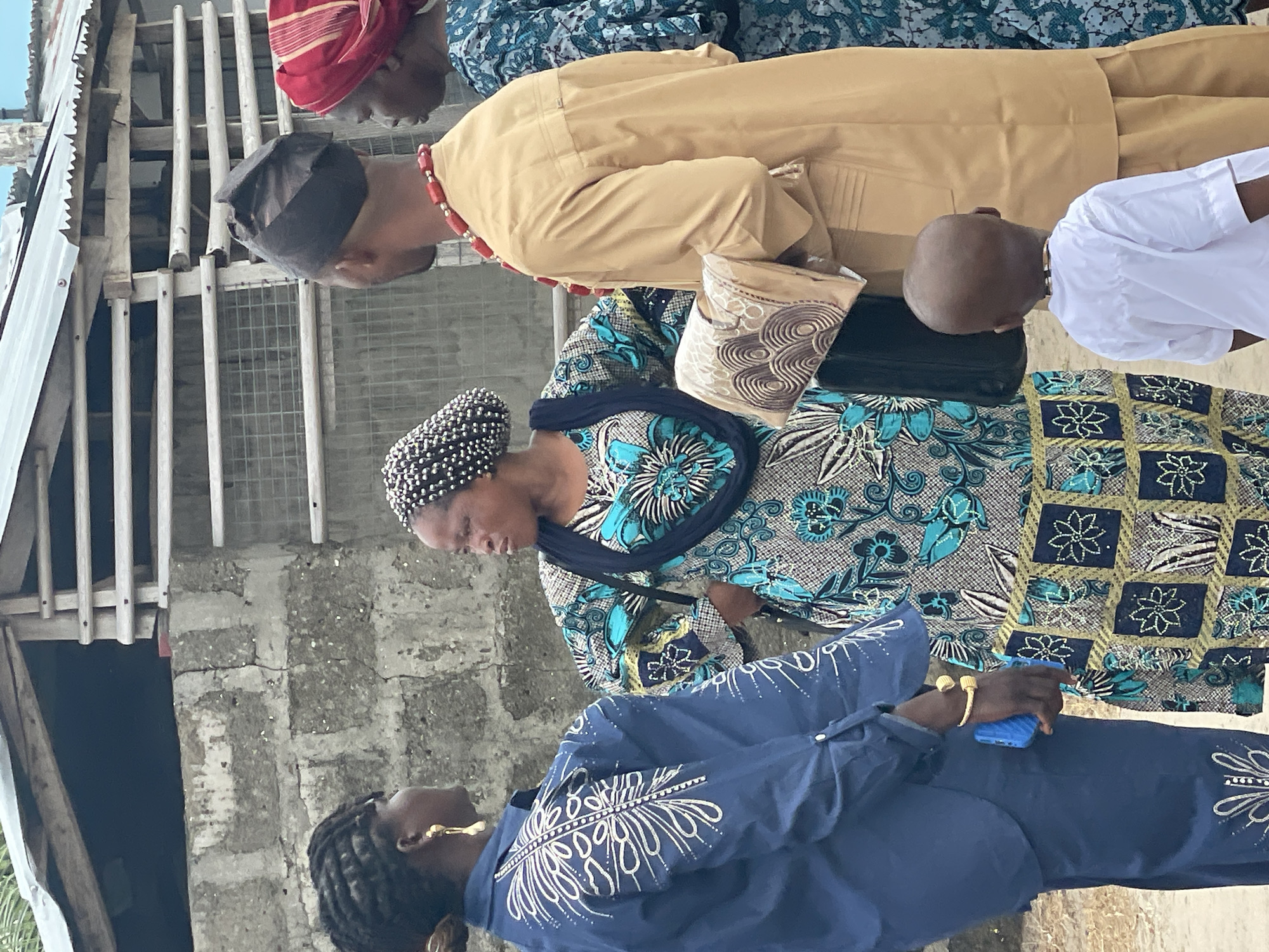 A group of people are gathered outdoors in Badagry, Nigeria, near a building with a corrugated metal roof and cinderblock walls. One woman in the foreground is wearing a long-sleeved blue dress with white embroidery and gold jewelry, holding a blue mobile phone. Behind her is another woman wearing a blue and gray patterned dress and a beaded head covering. A man in a tan tunic and red beaded necklace stands next to a boy in a white shirt, both looking in the same direction. The photo is taken at an angle, capturing the scene from a slightly elevated perspective.
