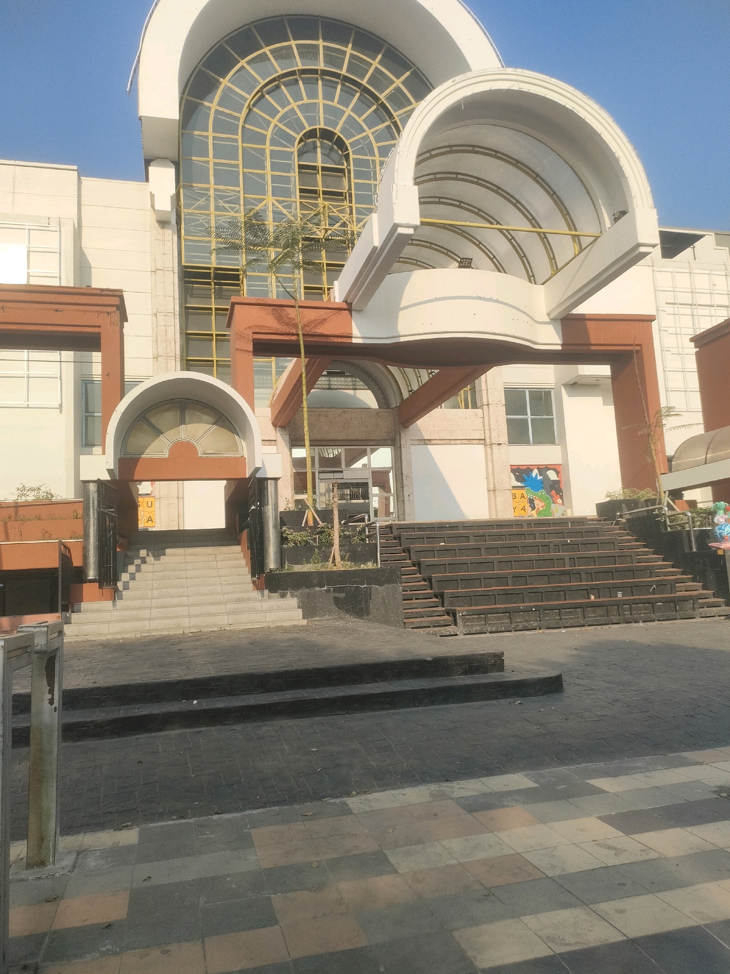 This is a full-shot, eye-level, outdoor photograph of the entrance to a modern building. The building has a large, arched, glass-paneled window on the upper level, with yellow framing. Below this, a white, arched canopy extends outward. The main entrance features a set of stairs leading up to a smaller arched doorway, flanked by dark metal railings. There are also wider, dark-colored tiered seating areas to the right of the stairs, leading up to another entrance. A mural depicting a cartoon character is visible on the wall behind the tiered seating. The ground in front of the building is paved with dark tiles. A young tree is planted near the entrance. The sky is clear and blue, suggesting a sunny day. The building appears to be located in Surabaya, Indonesia, based on the "BALAI KOTA SURABAYA" text visible on a yellow sign behind the stairs at the main entrance.