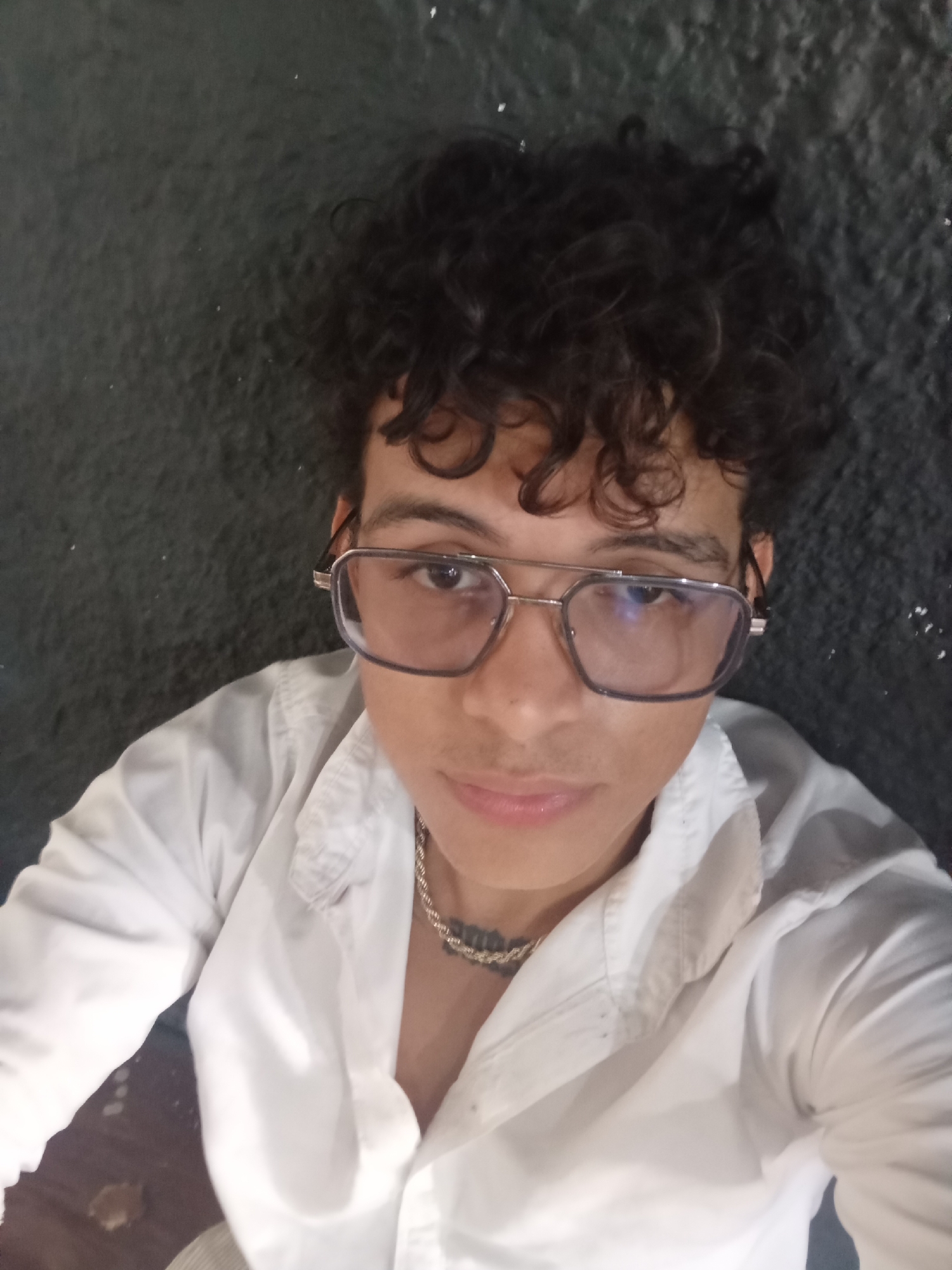 A medium close-up shot shows a person with curly dark hair and glasses, wearing a white collared shirt and a gold chain. The background is a dark, textured wall. The person's face is looking towards the camera, and they are wearing light-rimmed glasses. The shirt appears to be unbuttoned at the collar, revealing the gold chain. There is a subtle shadow cast on the left side of the image, suggesting the light source is to the right. The overall scene is dimly lit. The location in Juan Griego, Venezuela, is not discernible from the image itself.