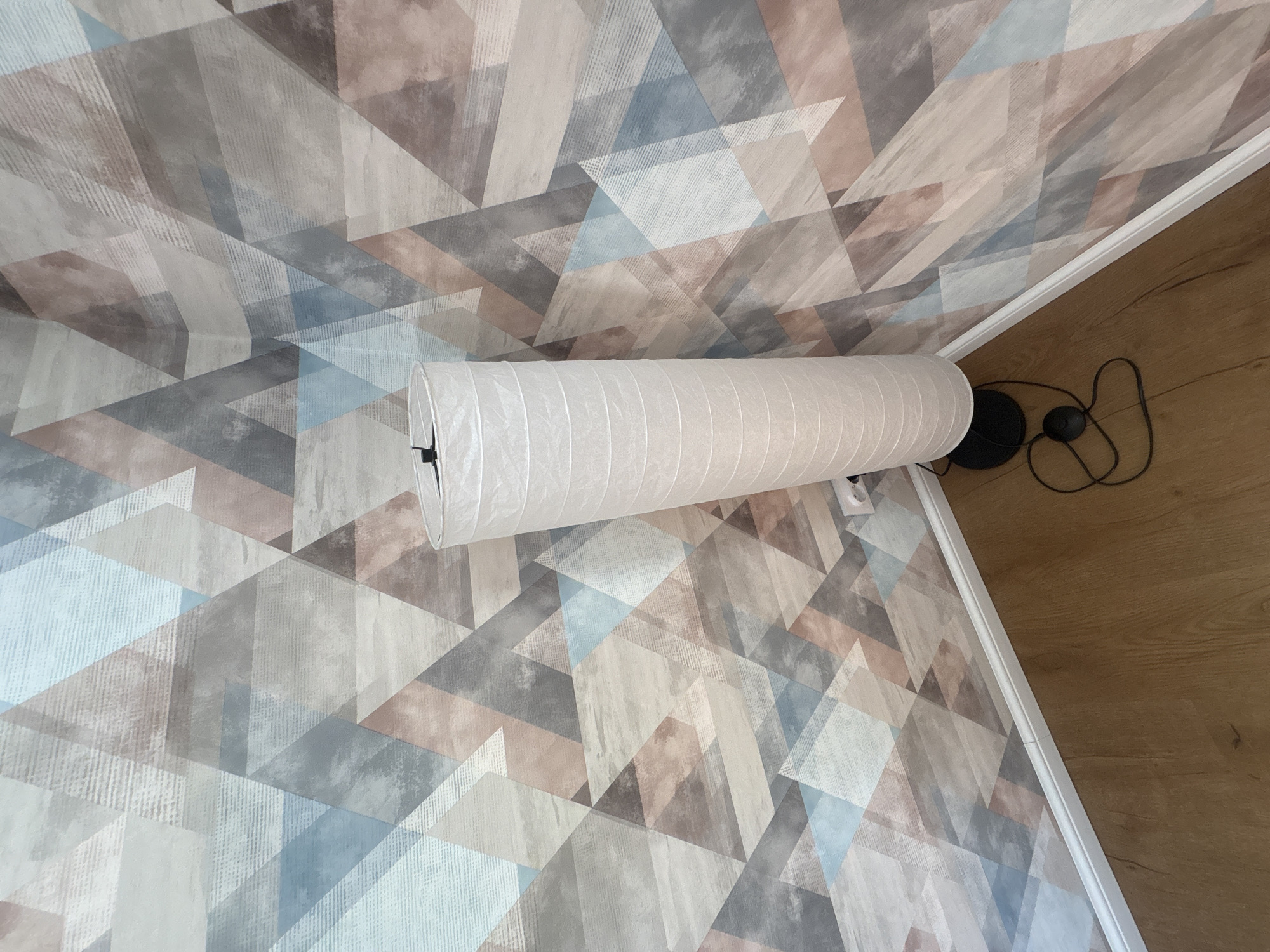 The image shows a view of a room corner. The walls are covered in geometric wallpaper with triangular shapes in shades of blue, gray, and brown. A long, cylindrical white lampshade is mounted horizontally on the wall. The floor is wooden, and there is a black power cord and a switch visible on the floor near the lamp.
