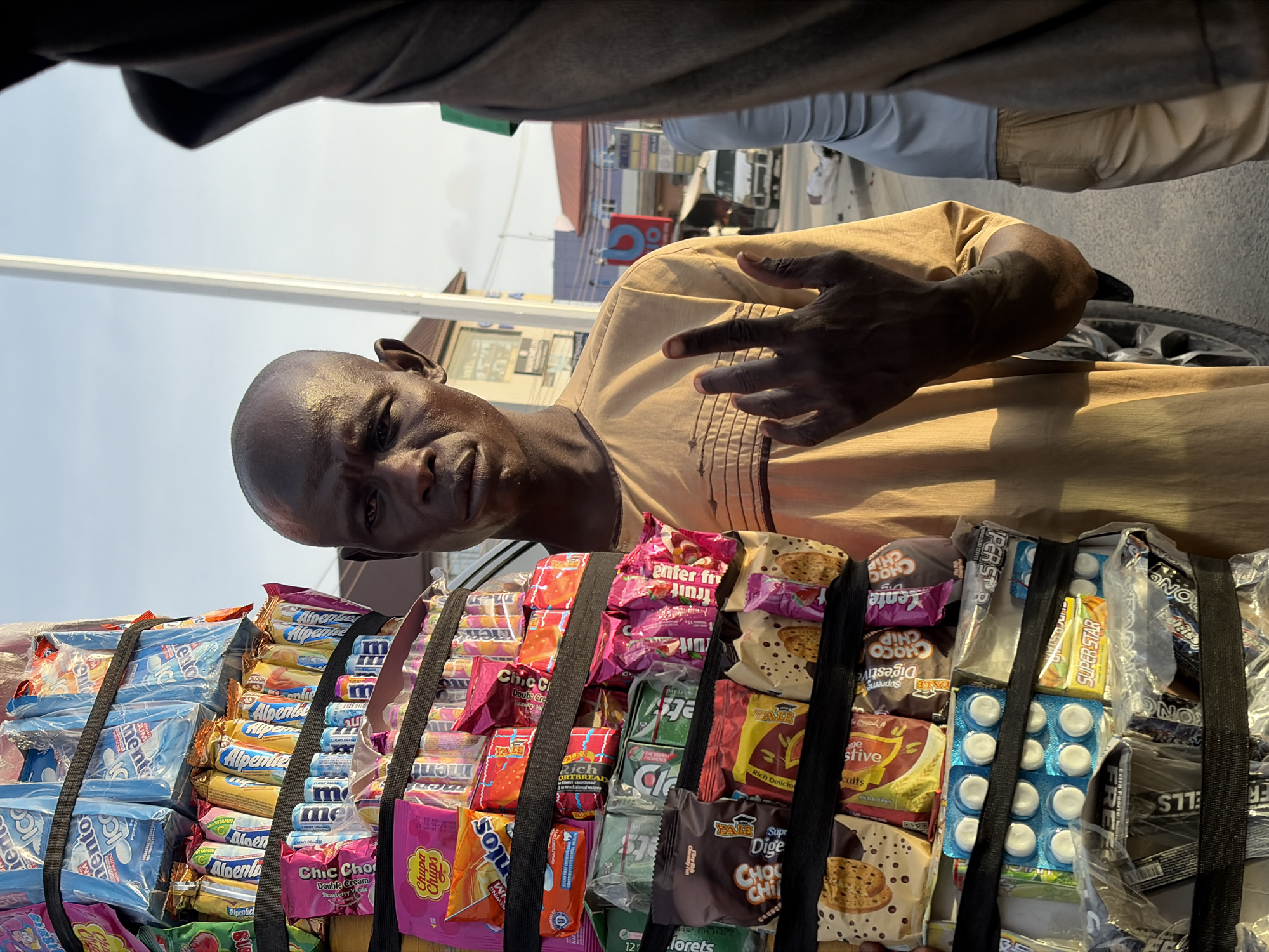 A mobile vendor in Ado, Nigeria, is displaying a variety of candy and snacks strapped together with black straps, likely for sale. The vendor, a Black man with a shaved head, is wearing a tan shirt with dark decorative stitching, looking directly at the camera with an expressionless face and holding his left hand up in a gesture. The assortment of treats includes brands like Mentos, Alpenliebe, Chic Choc, Chupa Chups, Yale, and others. The environment appears to be a city street, with other people and vehicles partially visible in the background.