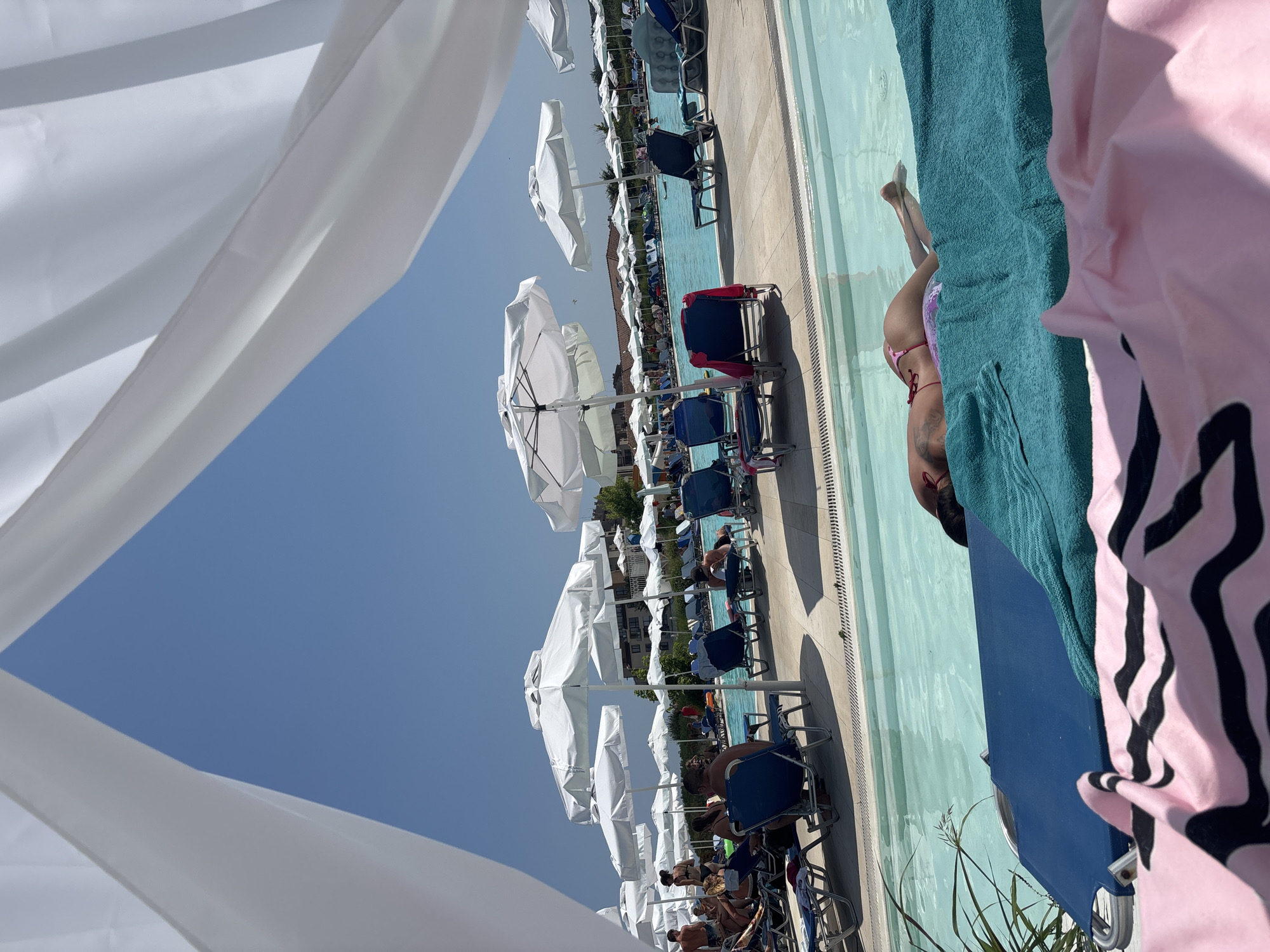 The image depicts a sun-drenched outdoor scene at a resort pool in Byala, Bulgaria. In the foreground, a person is lying on a blue lounger with a teal towel draped over them, partially obscured by a pink and black striped towel in the very front. The person is wearing a pink bikini and is positioned partially in the shallow end of the pool, with their legs extended.
The background shows a large, clear blue swimming pool surrounded by numerous white lounge chairs and white umbrellas. Several people are visible relaxing on the lounge chairs and walking around the pool area. In the distance, buildings that appear to be part of the resort complex are visible. The sky is a clear, bright blue. The scene is framed by white flowing fabric, likely a canopy or tent, in the upper part of the image. The overall impression is one of a typical summer day at a resort.