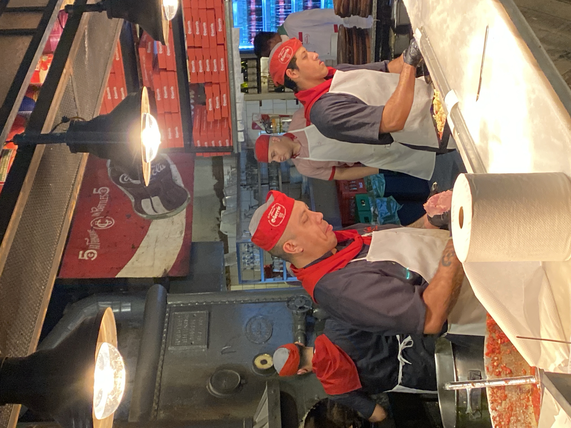 This image captures an indoor scene within a commercial kitchen or food preparation area, specifically Pizzería Güerrín, a prominent establishment located on Corrientes Avenue in Buenos Aires, Argentina, as evidenced by the "Güerrín" branding visible on staff caps and stacked red boxes.
Five adult males, all appearing to be staff members, are present and engaged in food preparation tasks. Four individuals in the foreground and mid-ground are uniformly dressed in red caps (some with "Güerrín" logos), red scarves, dark short-sleeved shirts, and white aprons. One man in the far background wears a white chef's jacket.
In the immediate foreground, a man with visible arm tattoos holds a piece of raw meat over a clean, white preparation counter, intently focused downwards. Adjacent to him on the right, another similarly uniformed and tattooed man, wearing black gloves, is actively assembling ingredients on a metallic tray. To the far left, a third uniformed individual is positioned over a large metal pot containing diced red ingredients, likely tomatoes. Behind this individual, a fourth uniformed man stands with his back partially to the camera, tending to a large, dark, industrial-style oven or cooking apparatus. In the background on the far right, the fifth individual in a white uniform stands near a refrigerated display case containing bottled beverages.
The environment is brightly lit by multiple industrial black pendant lamps suspended from the ceiling. The background features a large, vintage-style red and white sign with "5 BOTTLES 5" and a partially visible "Coca-Cola" logo on the left wall, and multiple shelves densely stacked with red-orange "Güerrín" branded boxes on the right. A roll of white paper towels and a small knife rest on the white countertop in the foreground. The scene depicts a busy, professional, and efficient food production setting.