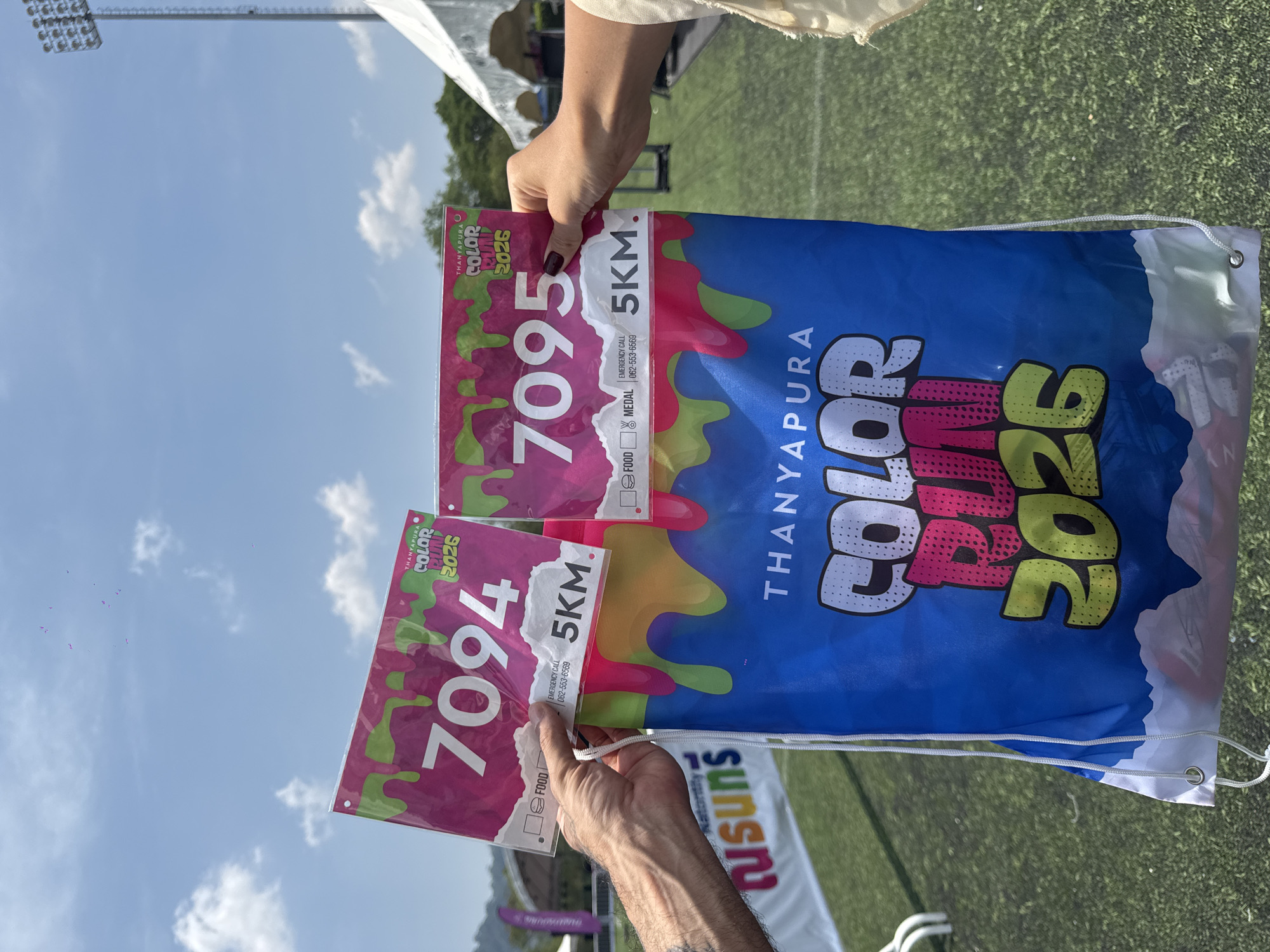 Two individuals are holding race bibs and a drawstring bag related to the Thanyapura Color Run 2026, in Thep Krasatti, Thailand. The bibs show the numbers 7094 and 7095, with the race distance listed as 5 km. The bag features the same event logo. The scene appears to be outdoors, evidenced by the presence of a field and light poles visible in the background. A small portion of a banner with the word "Tusuns" is also visible at the bottom of the image.
