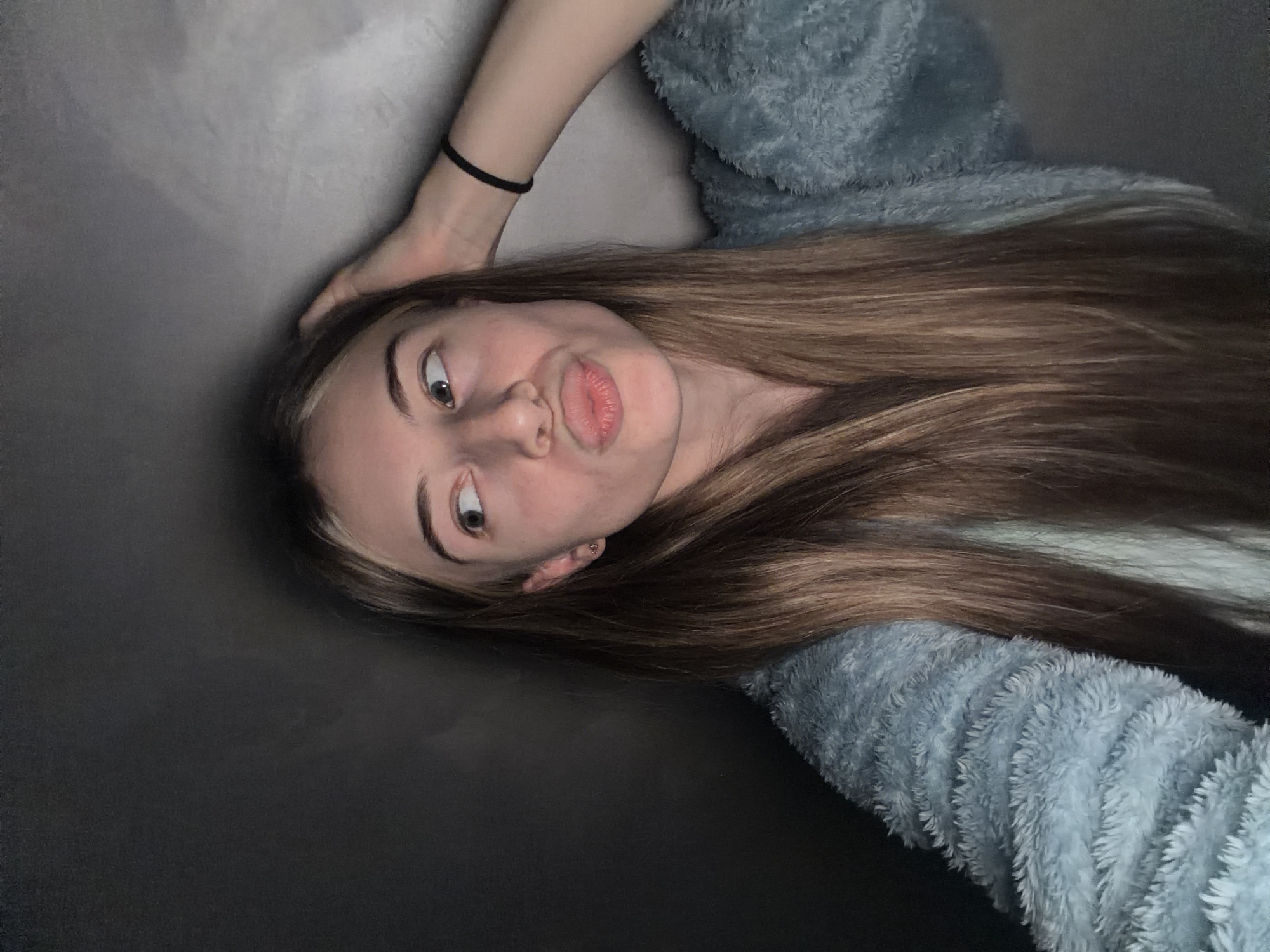 A young woman is lying down, presumably on a dark gray or black surface that could be a couch or bed. She is wearing a light blue, fuzzy-textured garment. Her long, light brown hair is spread out around her head. She is looking slightly to her left, with a slightly puckered expression on her face. A black elastic band is visible around her left wrist. An earring is visible in her left ear. No context indicates that the location is Plovdiv, Bulgaria.